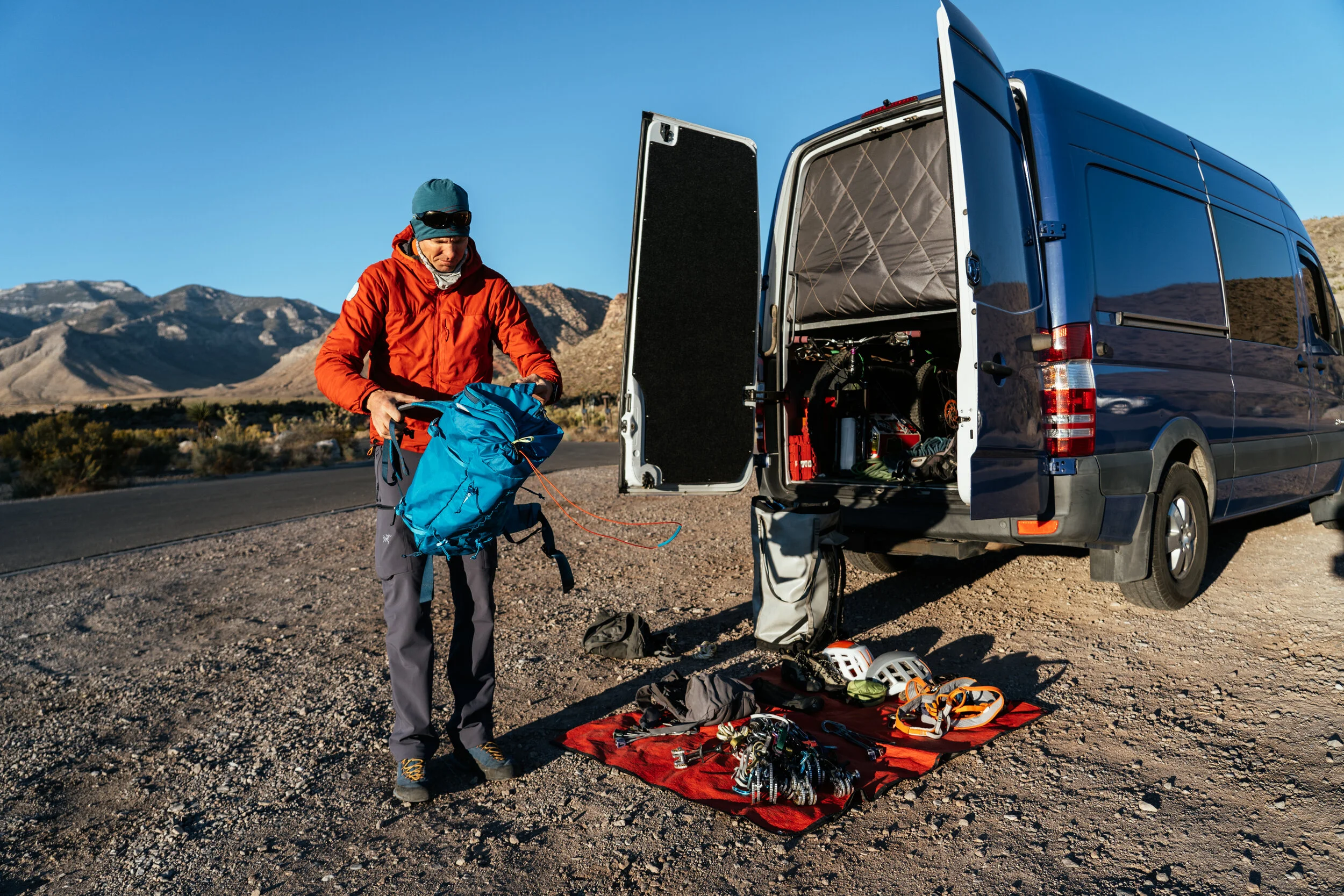 A person in outdoor mountain climbing gear standing beside a blue van with its rear doors open, with climbing equipment laid out on a red mat on the ground in a mountainous desert landscape.