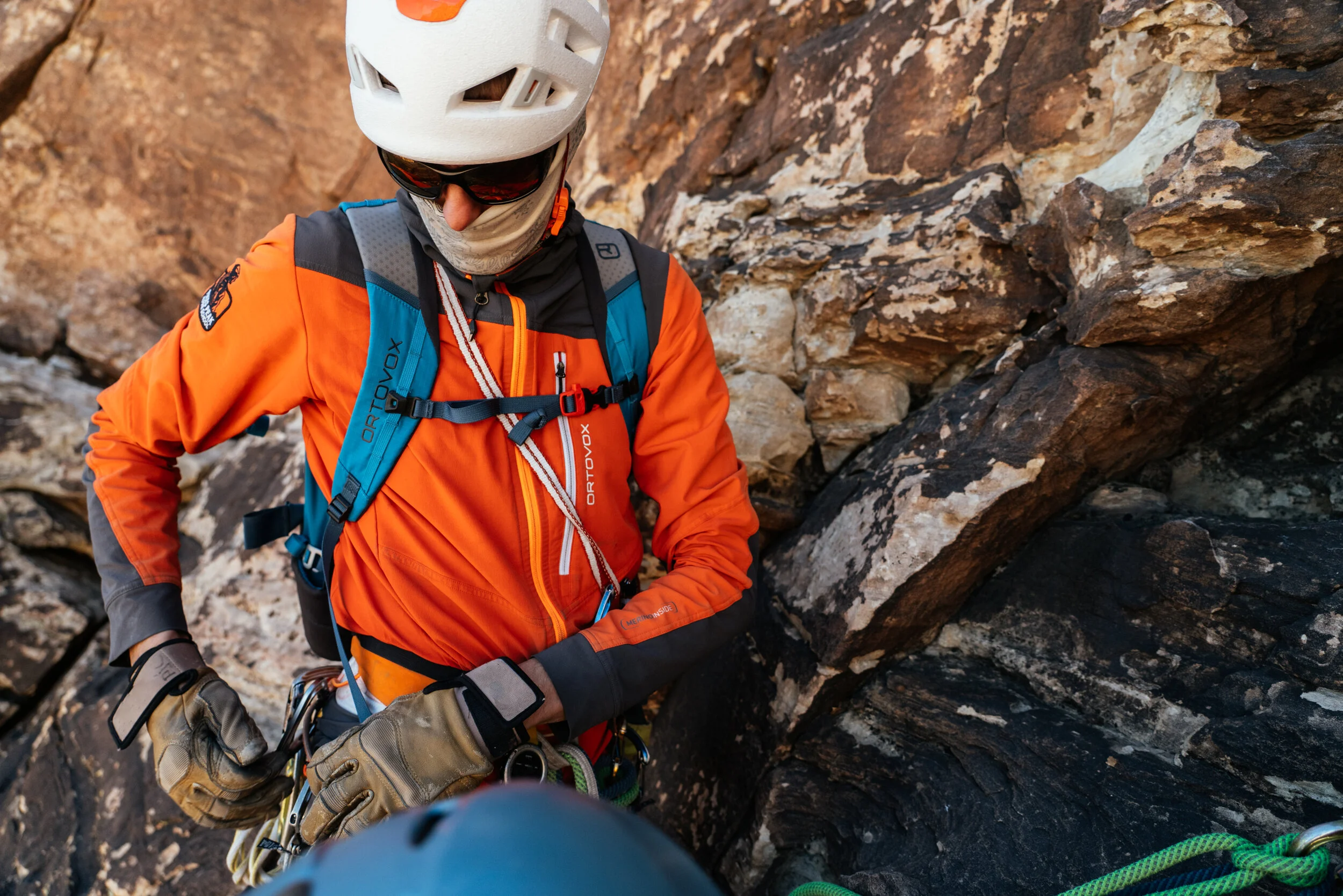 Climber with an orange jacket, white helmet, sunglasses, and gloves, securing climbing gear on a rock face.