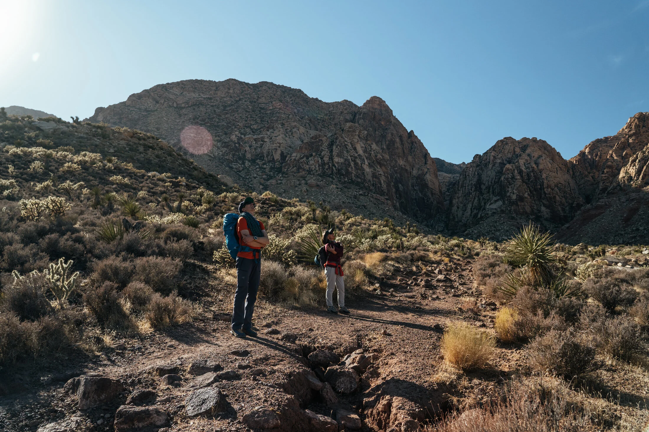 Two hikers standing on a rugged trail in a desert landscape with mountains in the background, under a bright blue sky.