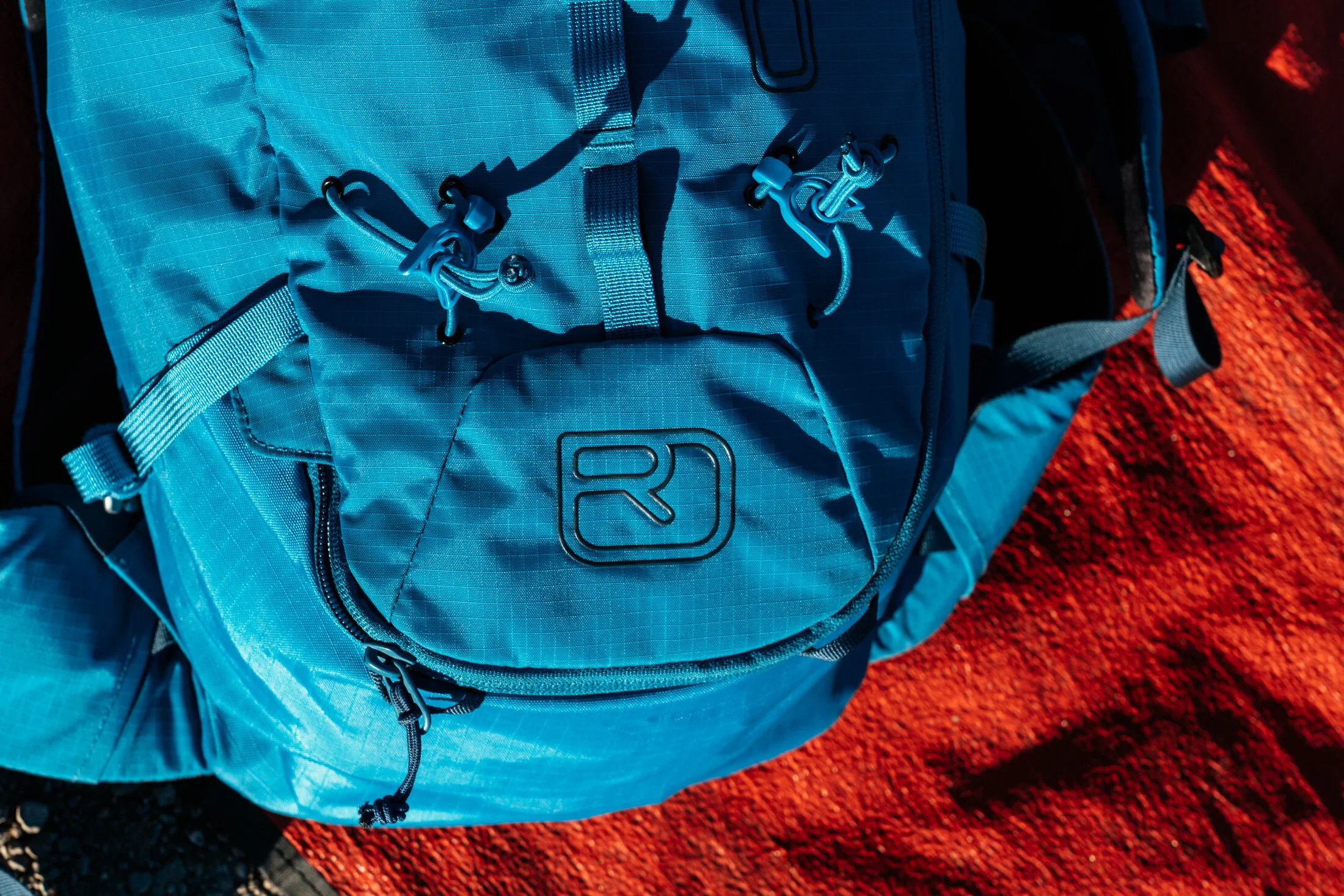 Close-up of a blue outdoor backpack with adjustable straps and multiple compartments, placed on a reddish ground.