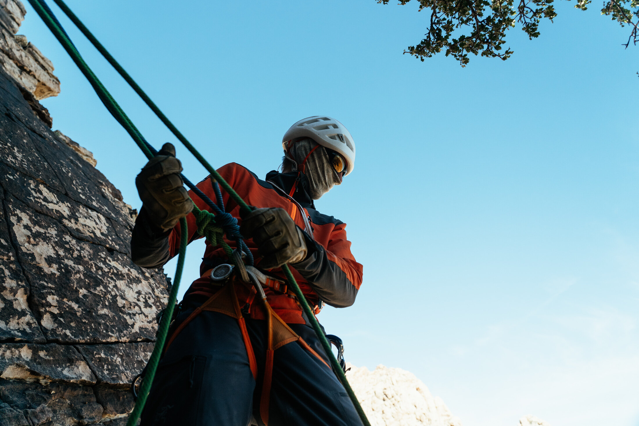 Climber wearing a helmet, sunglasses, and a face covering, adjusting ropes on a rocky cliff with a clear blue sky above.