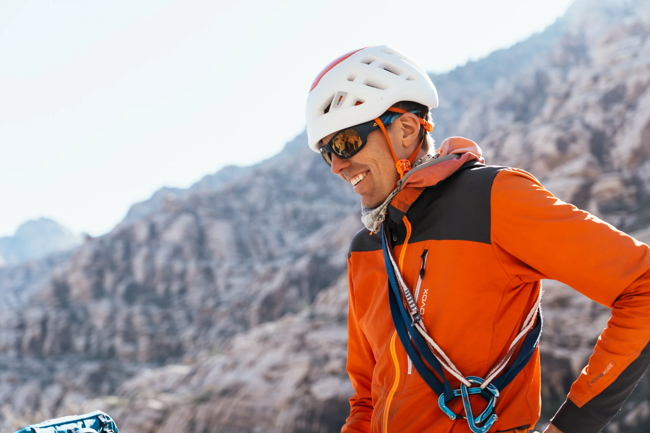 Smiling man wearing an orange jacket, white helmet, and sunglasses outdoors with a rocky mountain background.