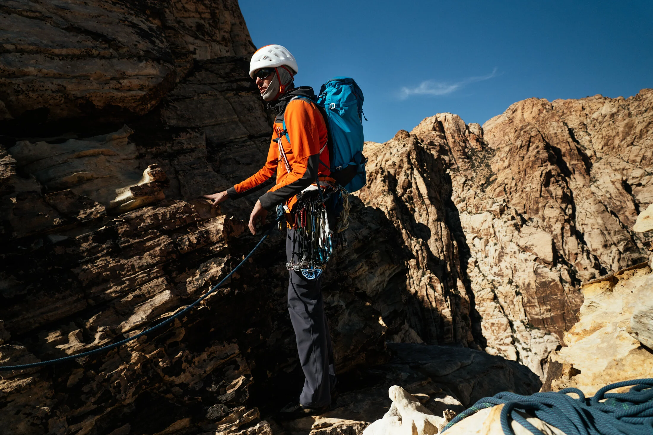 A person in orange and black climbing gear, helmet, and sunglasses climbs rocky terrain in a mountainous area with visible cliffs and clear blue sky.