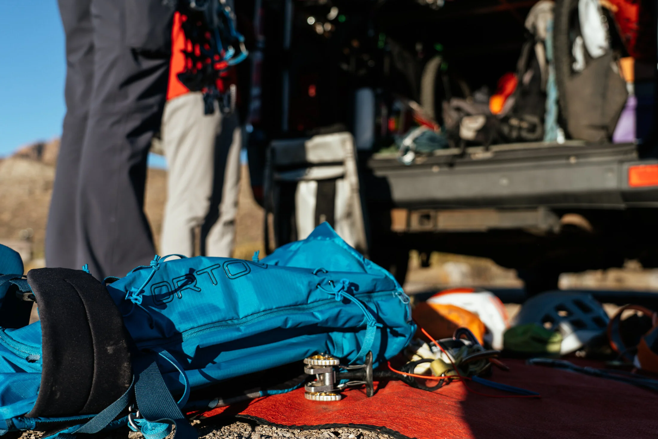 Climbing gear and equipment laid out on the ground in front of an open vehicle, with a person standing in the background.