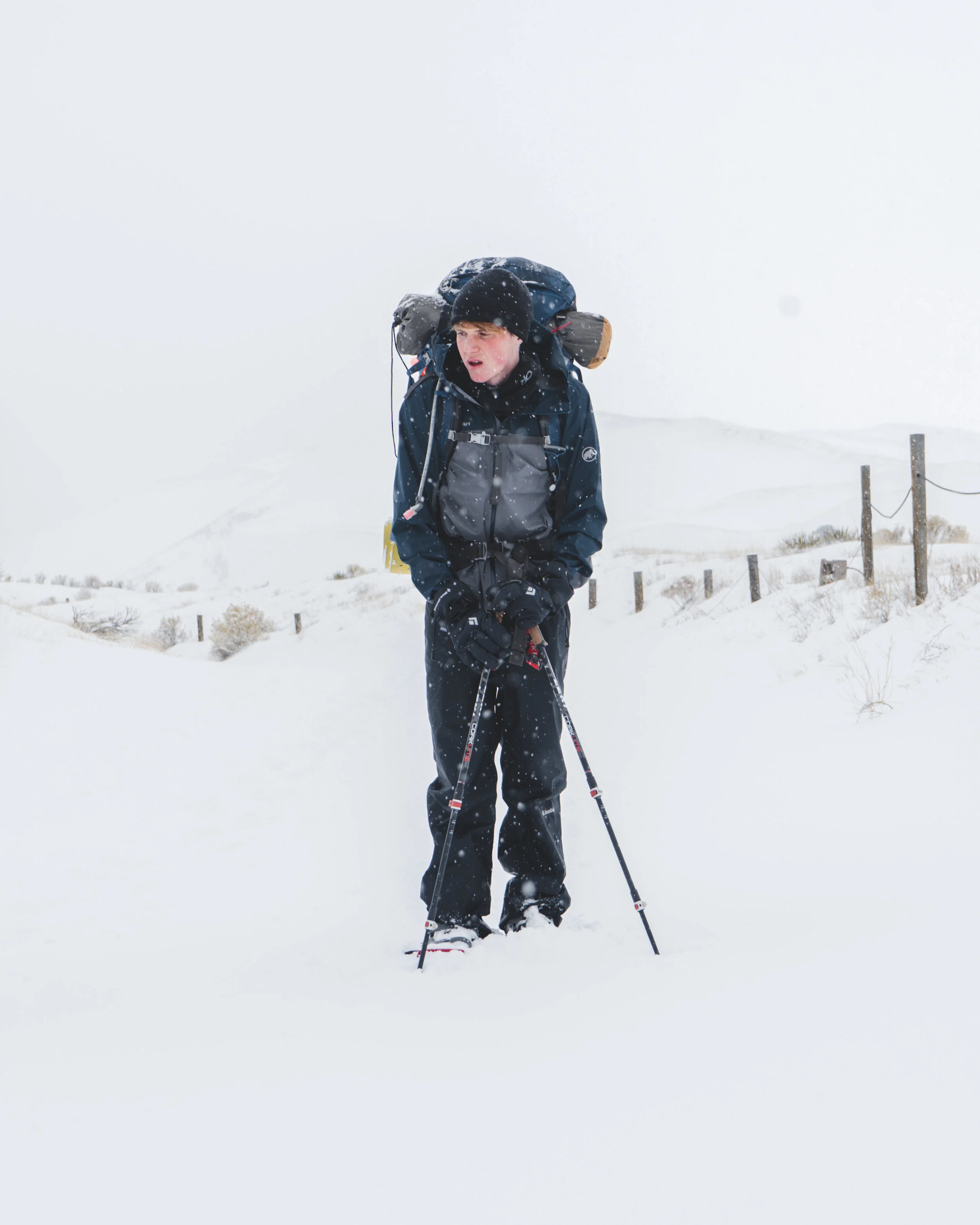 A person dressed in winter gear, carrying a large backpack, stands in a snowy landscape with snow falling, holding ski poles, and appears to be snowboarding or snow trekking.