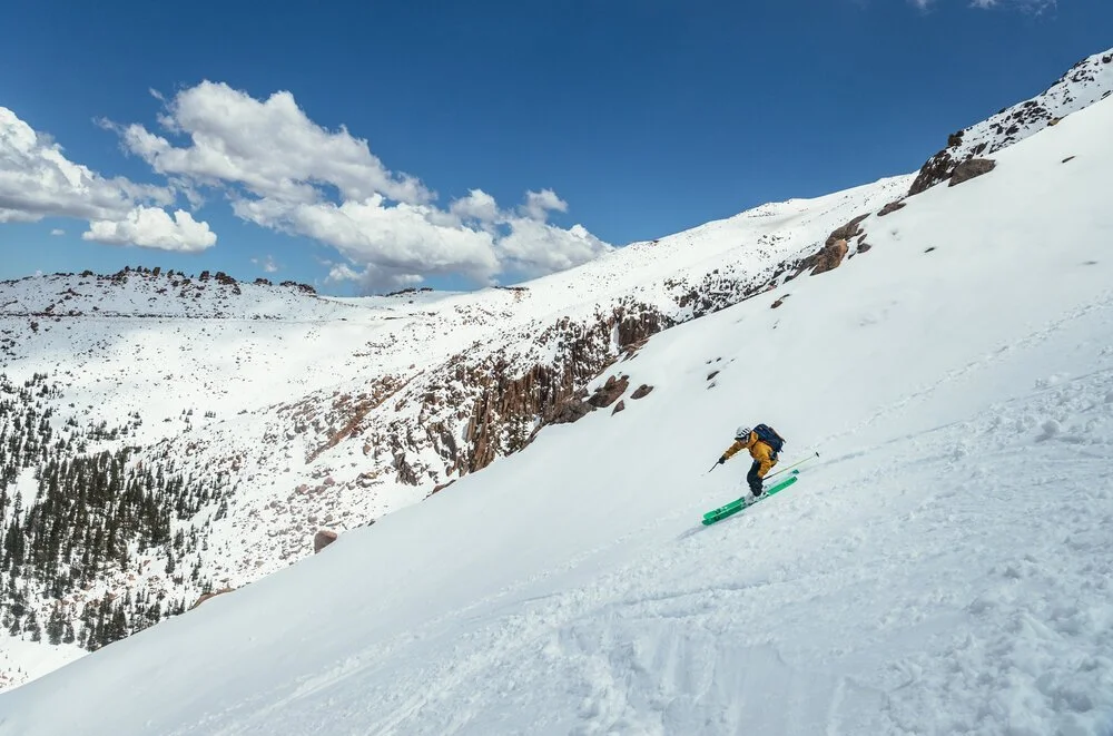 A person skiing down a snowy mountain slope with a backpack, under a blue sky with clouds, in a winter landscape of snow-covered mountains and pine trees.