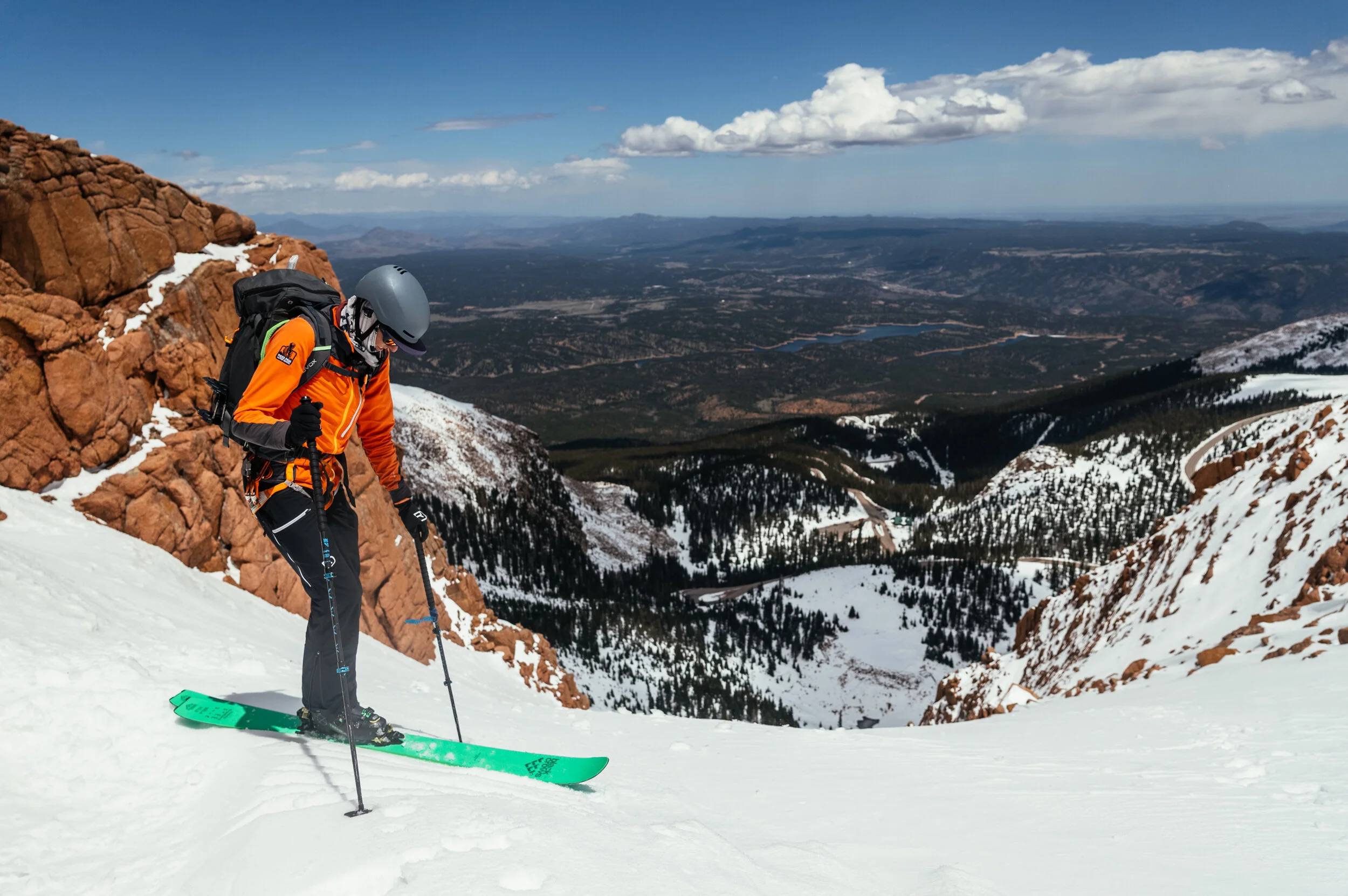 A skier in an orange jacket and black pants skiing on snowy mountain with a scenic view of valleys, forests, and a lake in the distance.