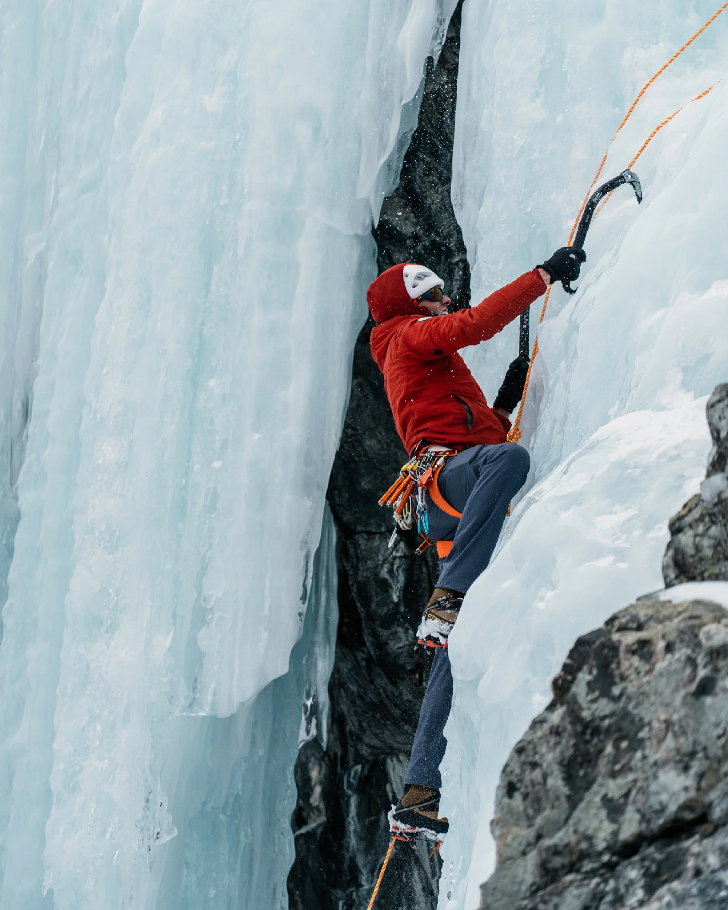 A person ice climbing on a frozen glacier or ice wall, wearing a red jacket, helmet, and climbing gear, with ice axes and crampons.