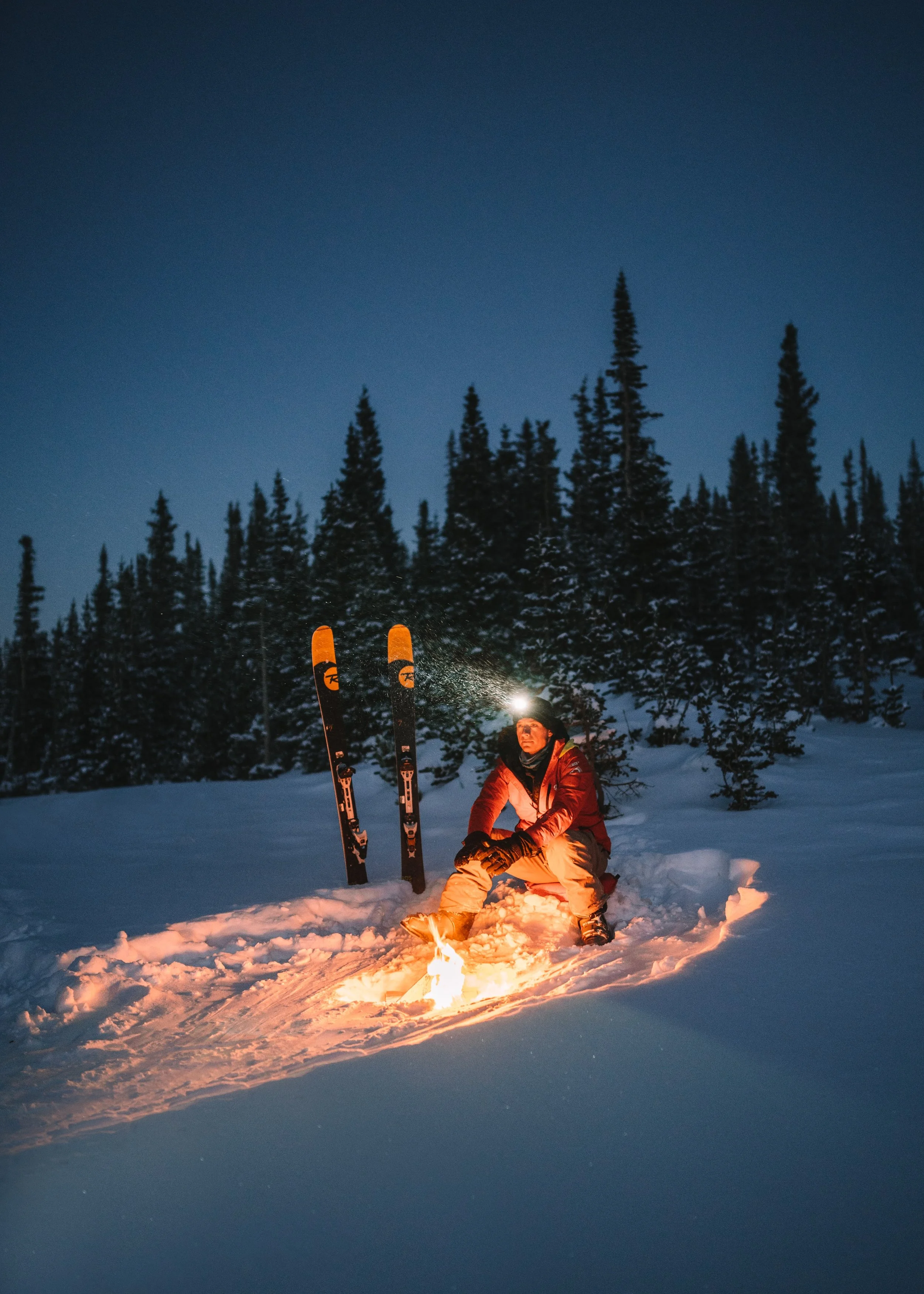 A person wearing winter gear sits by a small fire in a snowy landscape at night, with tall pine trees in the background, and skis standing upright in the snow beside them.