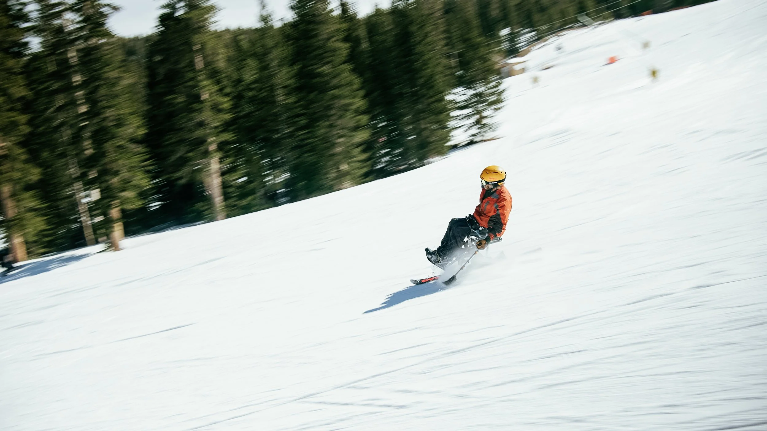 A person skiing downhill on a snow-covered slope with trees in the background, wearing a yellow helmet and an orange and black jacket.