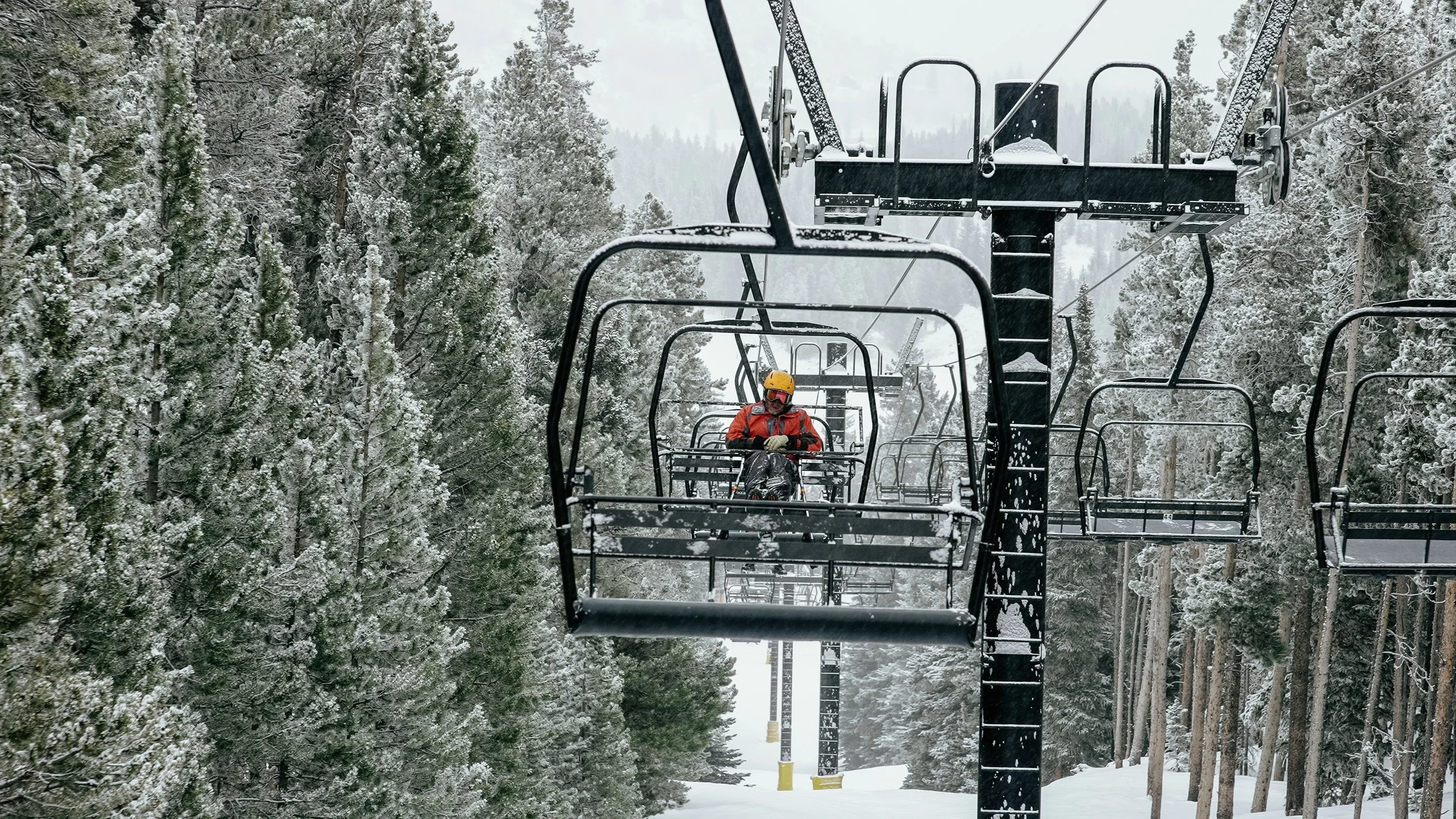 A person wearing an orange jacket, yellow helmet, and goggles riding a ski lift over a snowy forest with snow-covered trees.