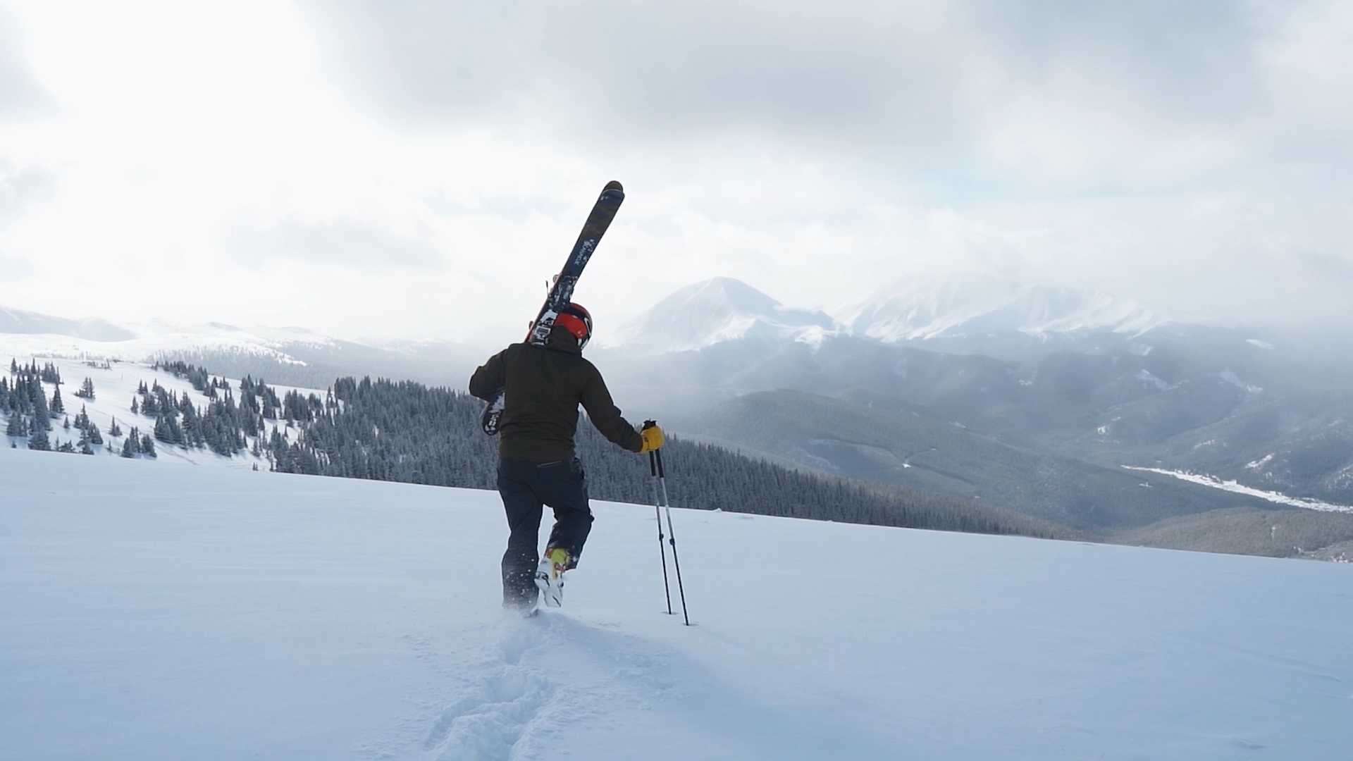 Person carrying skis and walking through snow on a mountain with a cloudy sky and distant mountains.