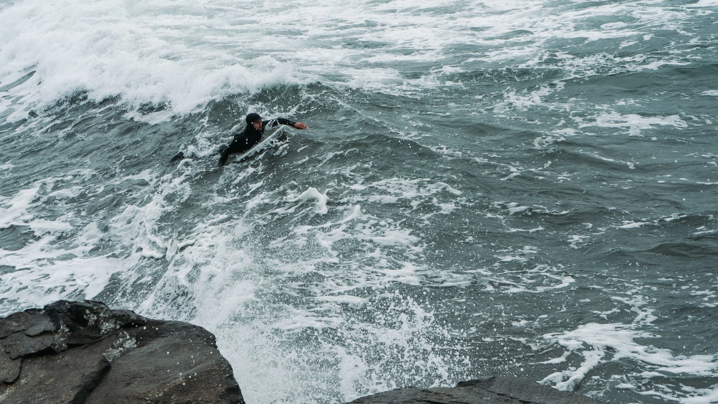 A person wearing a wetsuit and helmet is swimming away from rocks in rough ocean water, possibly after falling off a surfboard or boat.