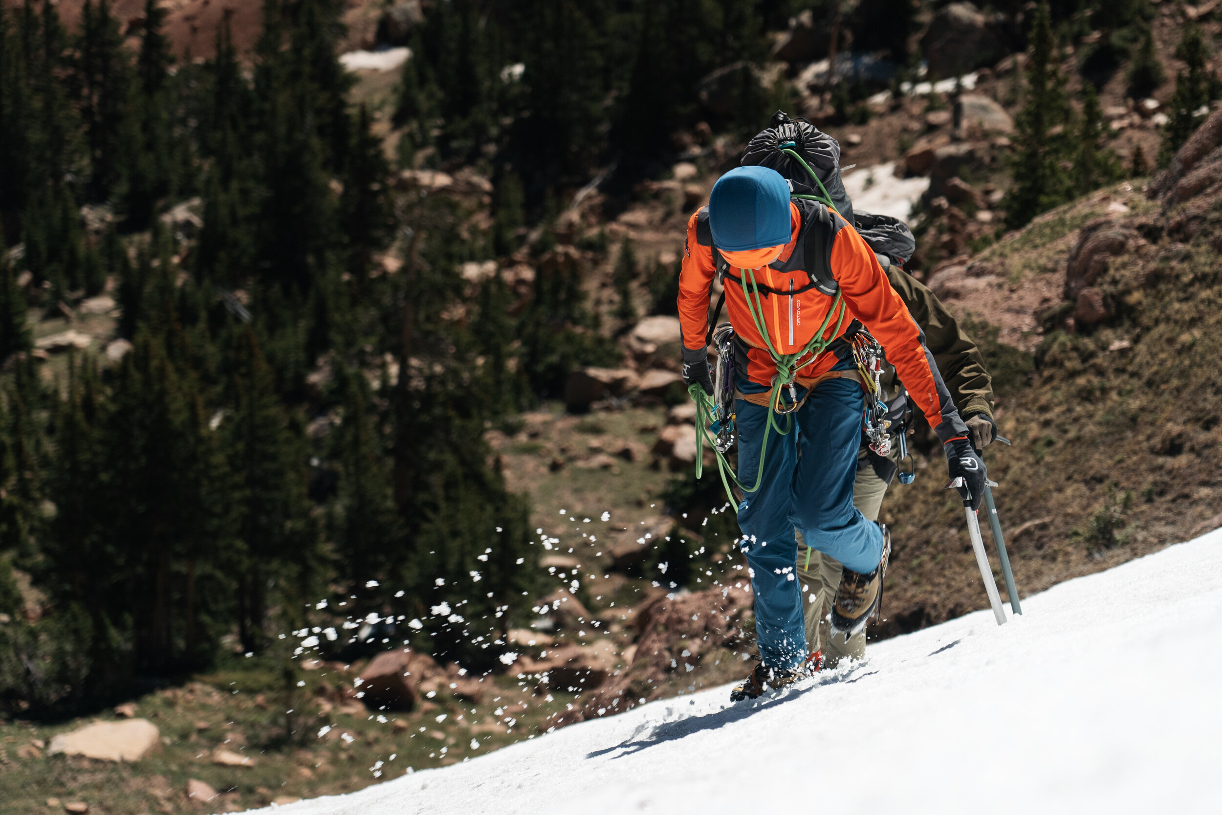 Two climbers ascending a snowy mountain slope, one in the foreground wearing an orange jacket, blue pants, and a blue helmet, with climbing gear and ice axes, and the other partially visible behind, also equipped for snow climbing. A forested mountai