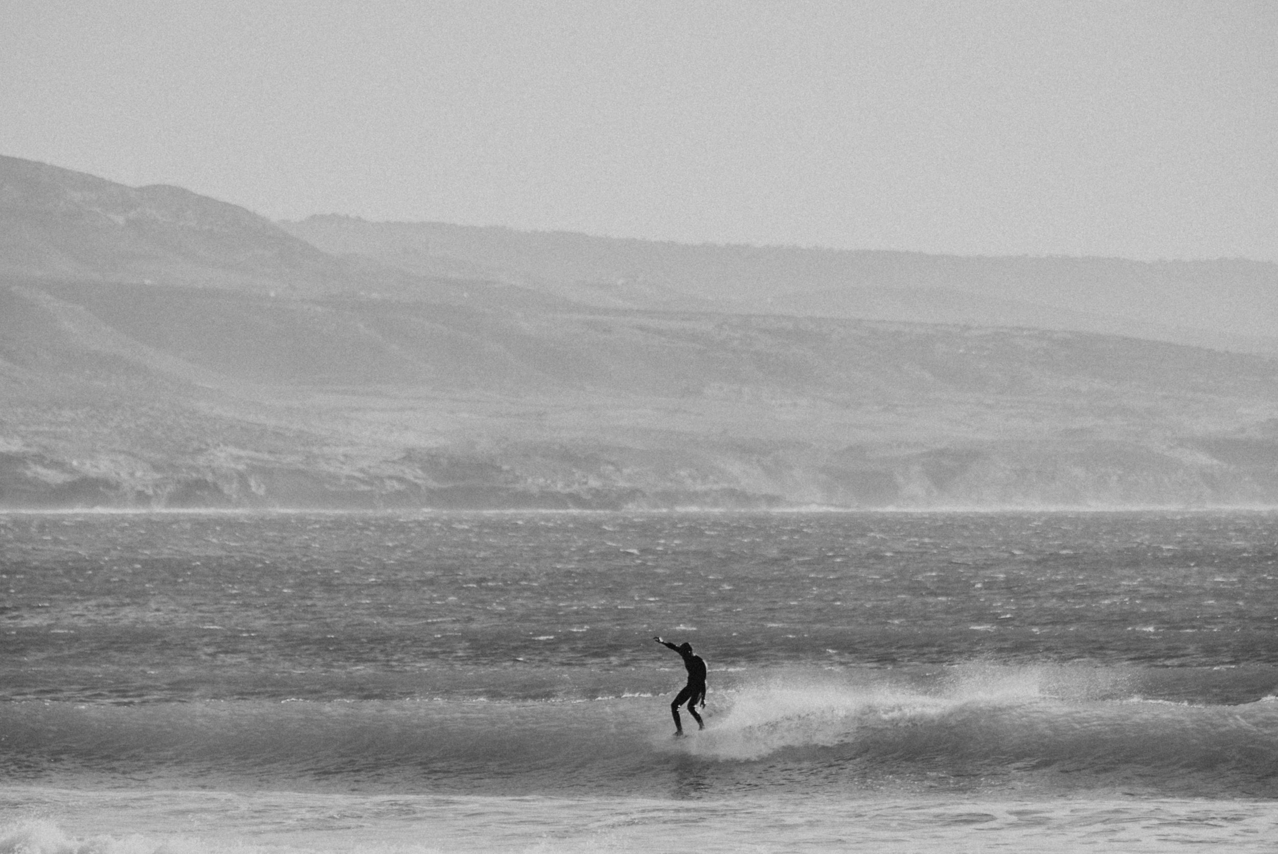 A person surfing on a wave in the ocean with distant hills in the background, in black and white.