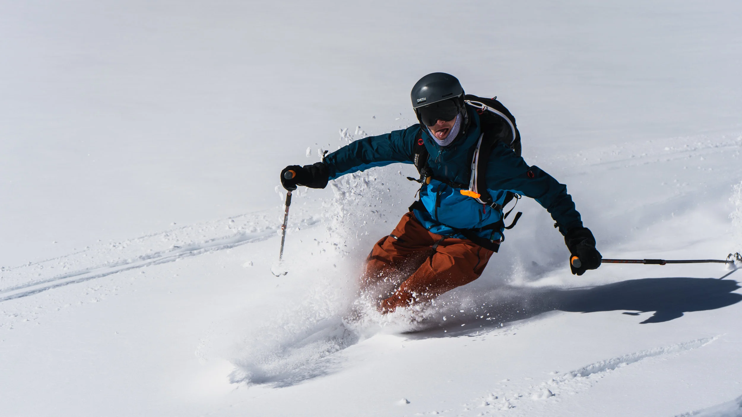 Person skiing down a snowy slope wearing a helmet and goggles, dressed in a blue jacket and brown pants, creating a spray of snow as they carve through the snow.