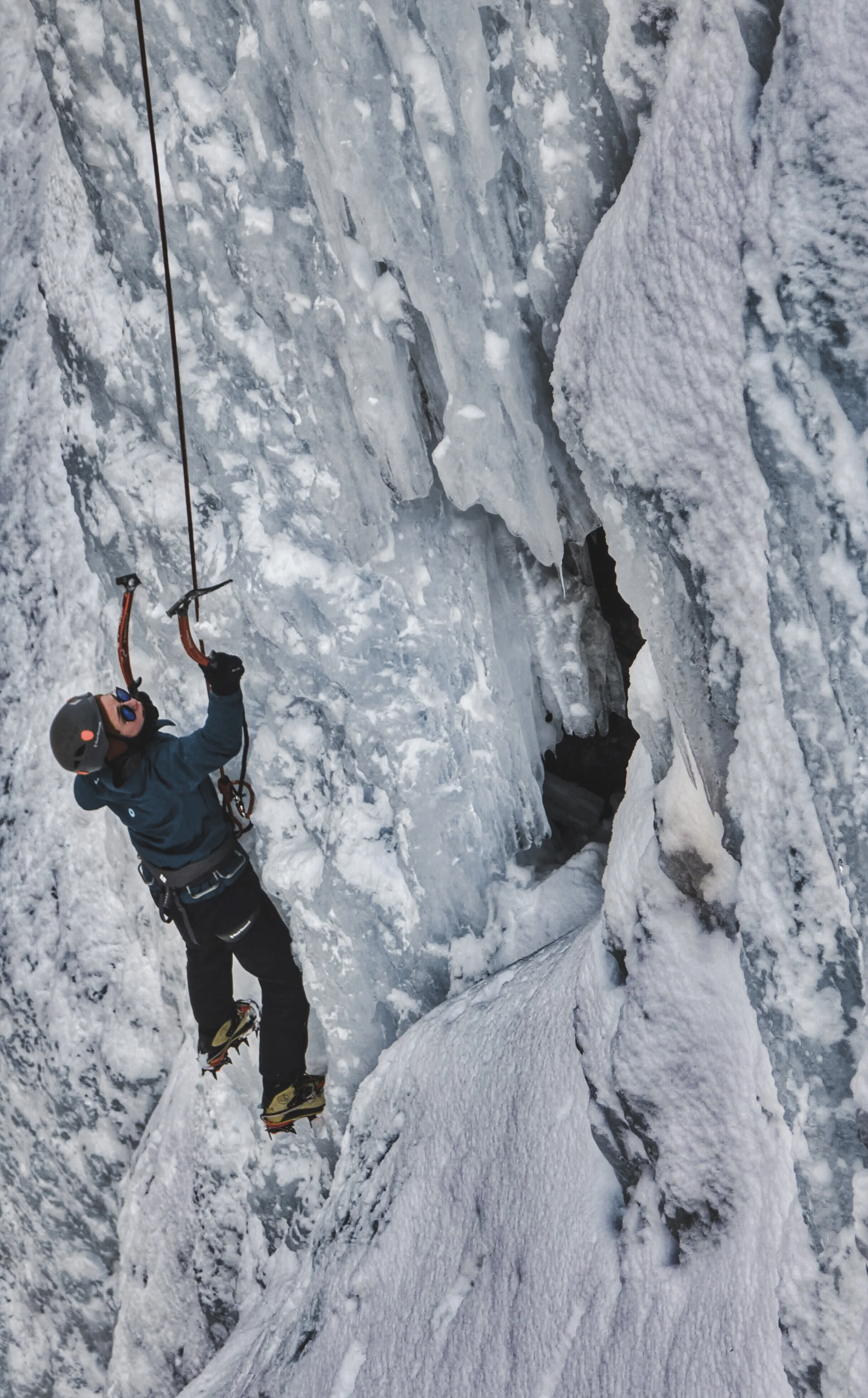A person ice climbing on a frozen wall with ice tools and crampons, wearing a helmet and winter gear.