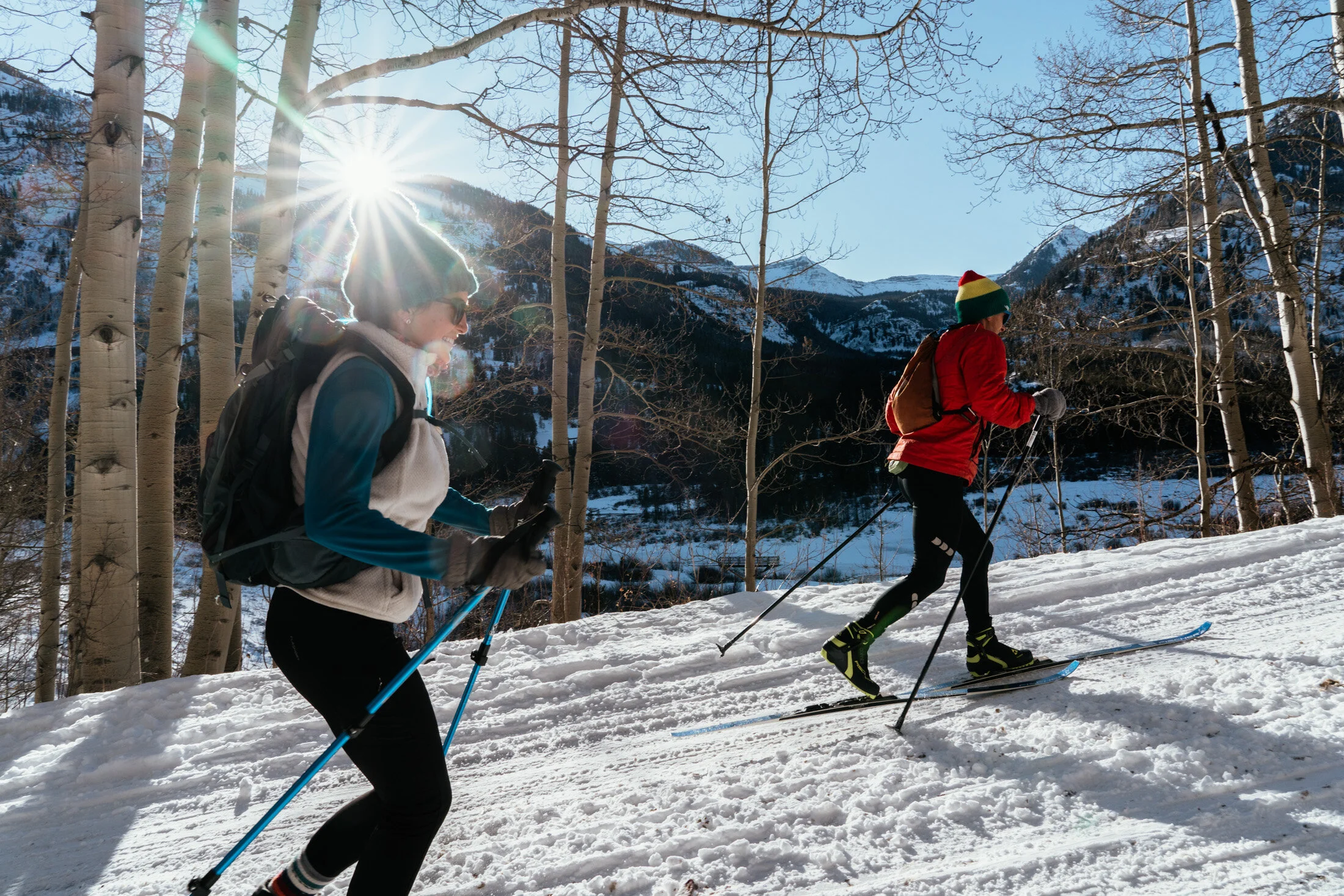 Two people cross-country skiing on a snowy trail in a forest with mountain background, sunlight shining through trees.