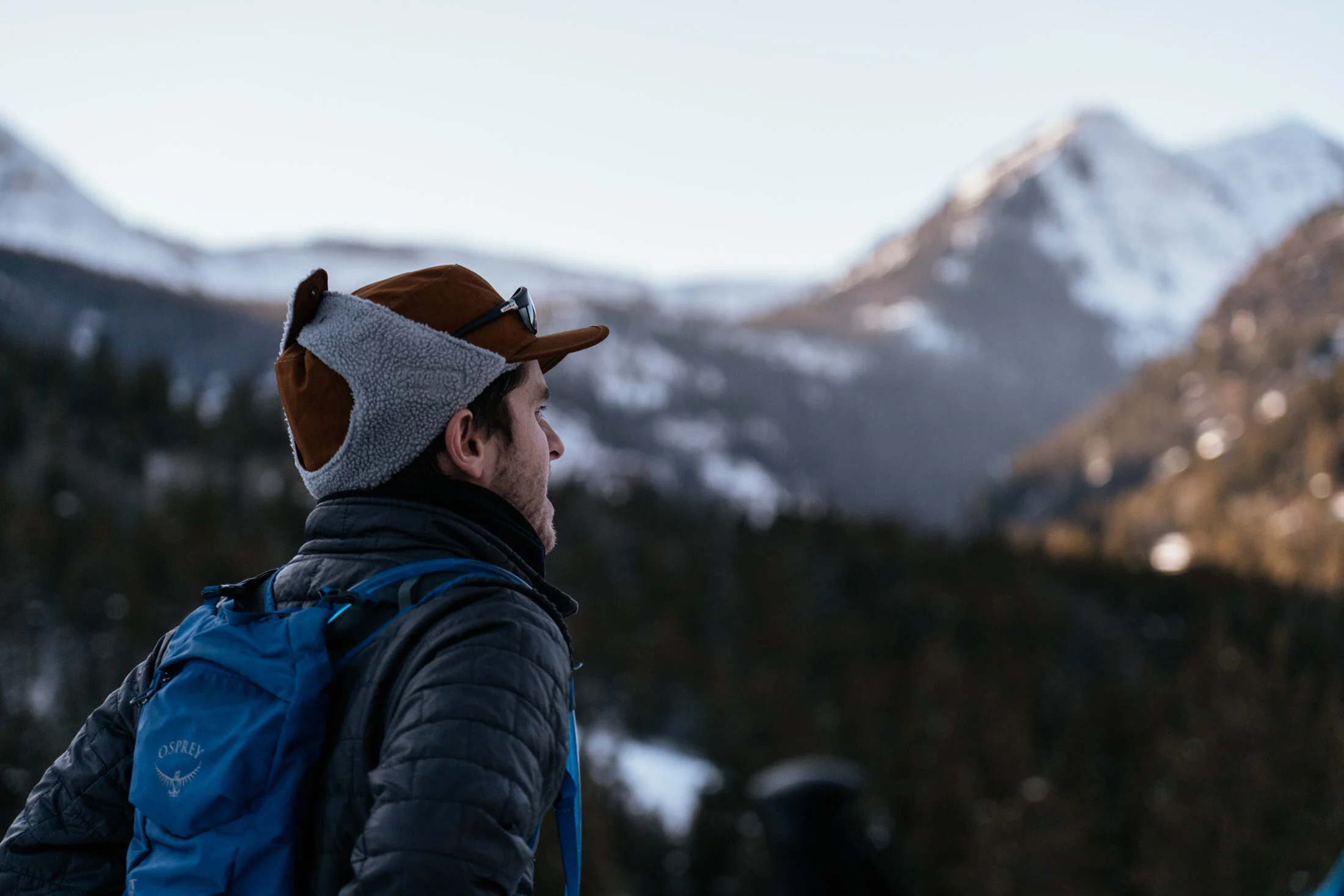 A male hiker wearing a brown cap, fall hoodie, and black jacket with a blue backpack, standing outdoors in a mountainous area with snow-covered peaks in the background.
