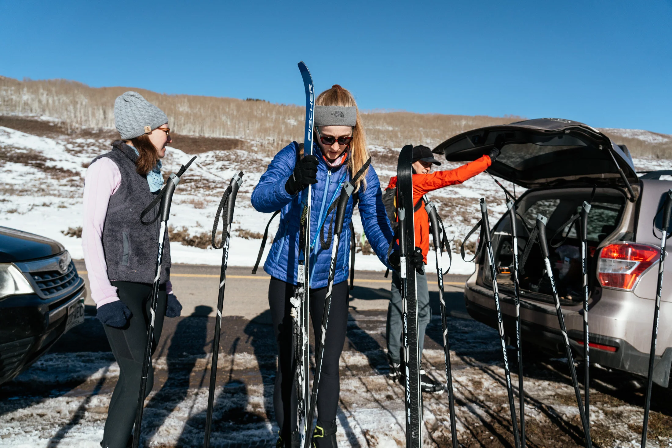 Three women preparing for a day of cross-country skiing near the car, which has its trunk open, on a snowy road with snow-covered hills in the background.