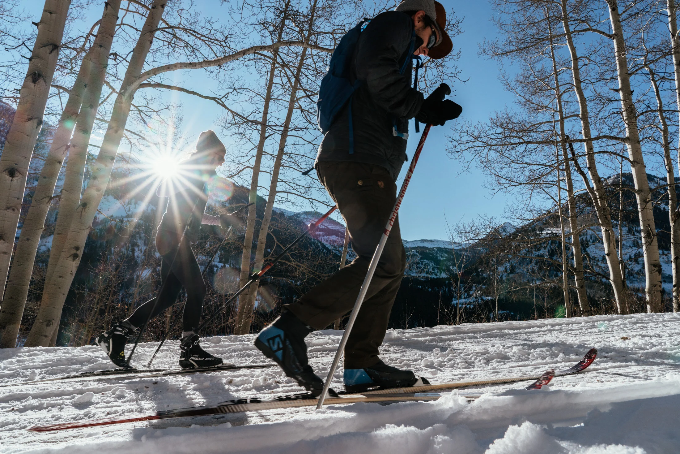 Two people cross-country skiing on a snow-covered trail surrounded by bare trees, with mountains in the background and the sun shining through the trees.
