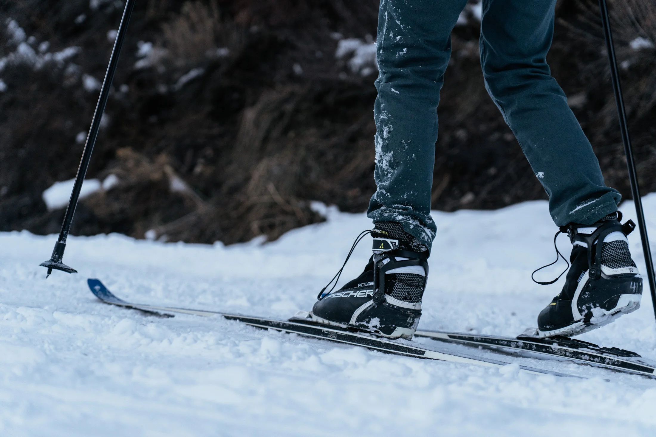Close-up of a person wearing ski boots and pants, standing on skis on snow with ski poles in hand.