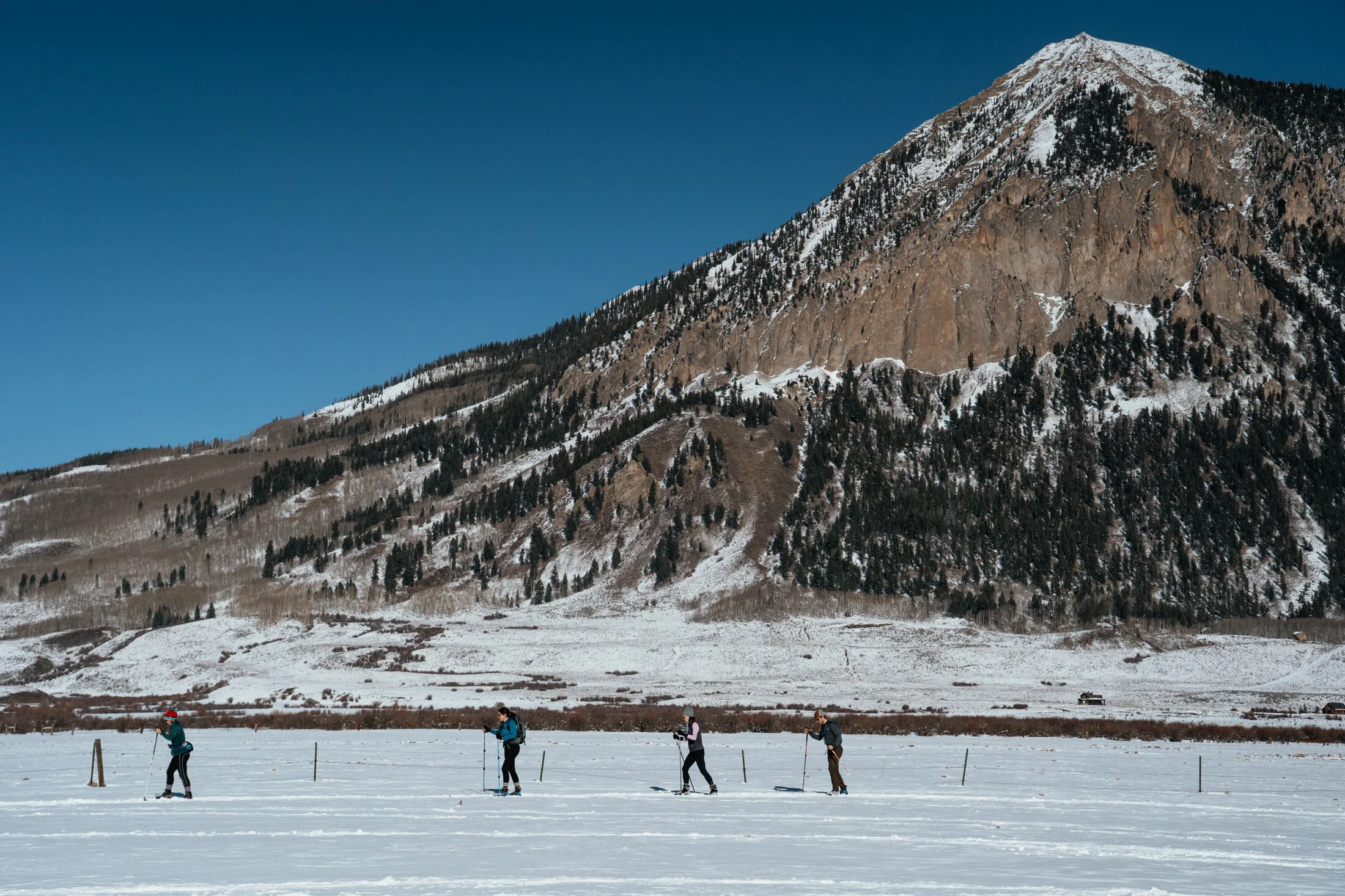 Four people cross-country skiing on a snowy field with a mountain backdrop under a clear blue sky.