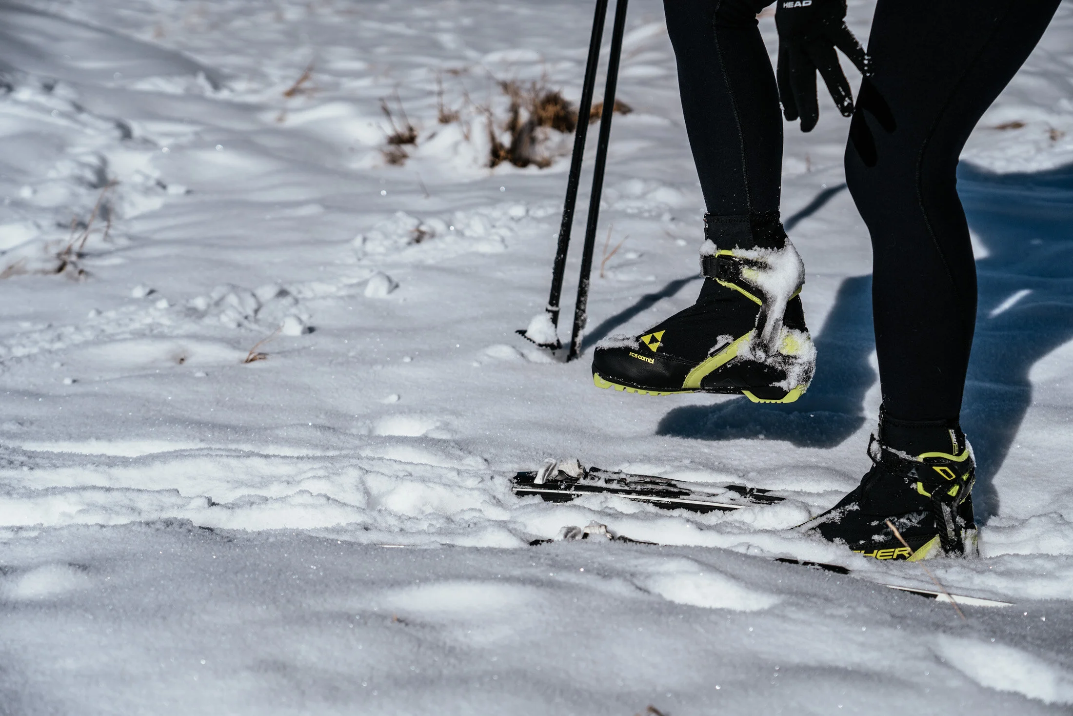 Person wearing snow boots and gloves standing on snow with ski poles, preparing to cross-country ski in a snowy landscape.