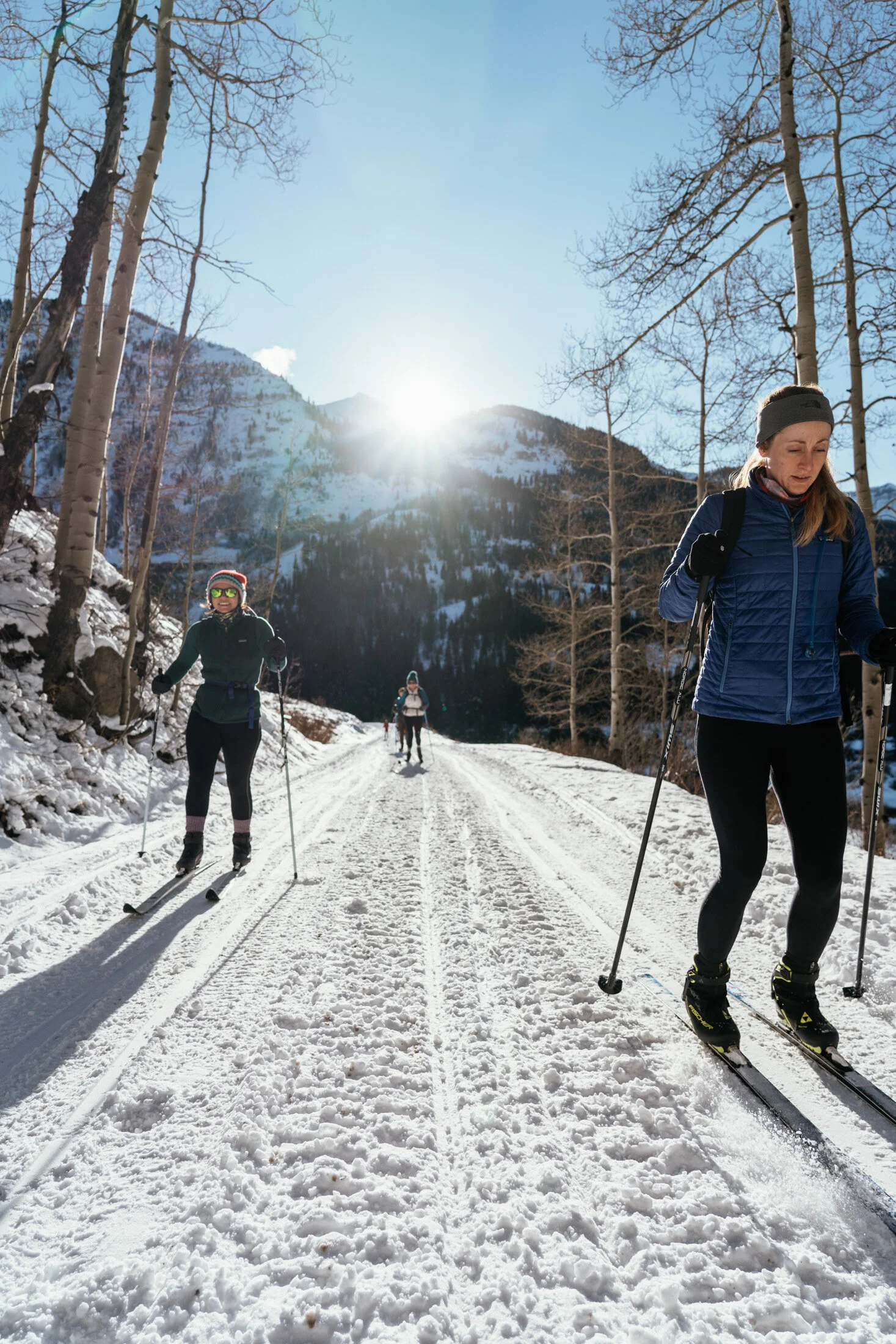 Three women cross-country skiing on a snowy trail in a mountainous area under a clear blue sky, with the sun shining brightly and leafless trees along the path.