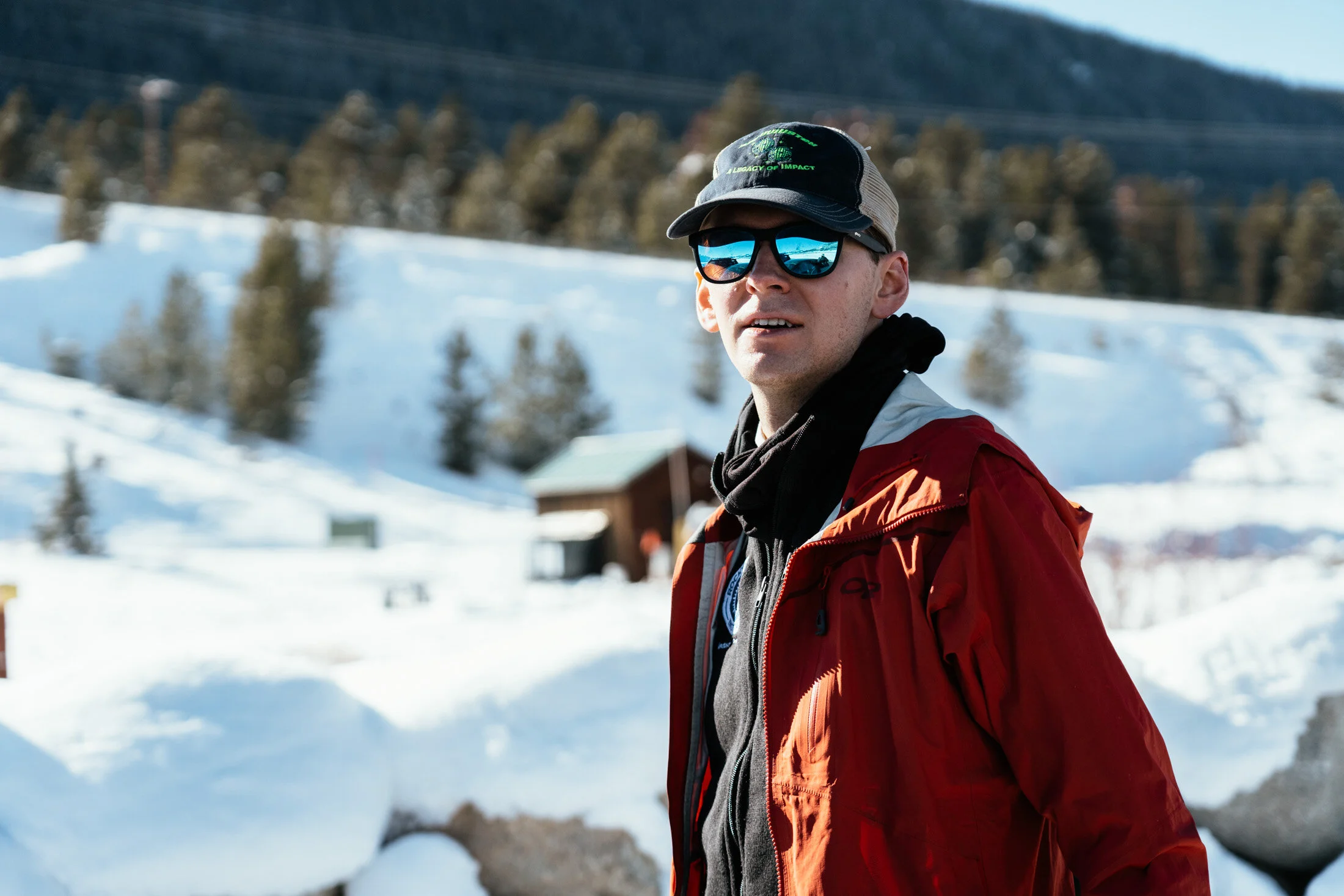 A man wearing sunglasses, a black cap, and a red jacket outdoors in a snowy landscape with trees and small buildings in the background.