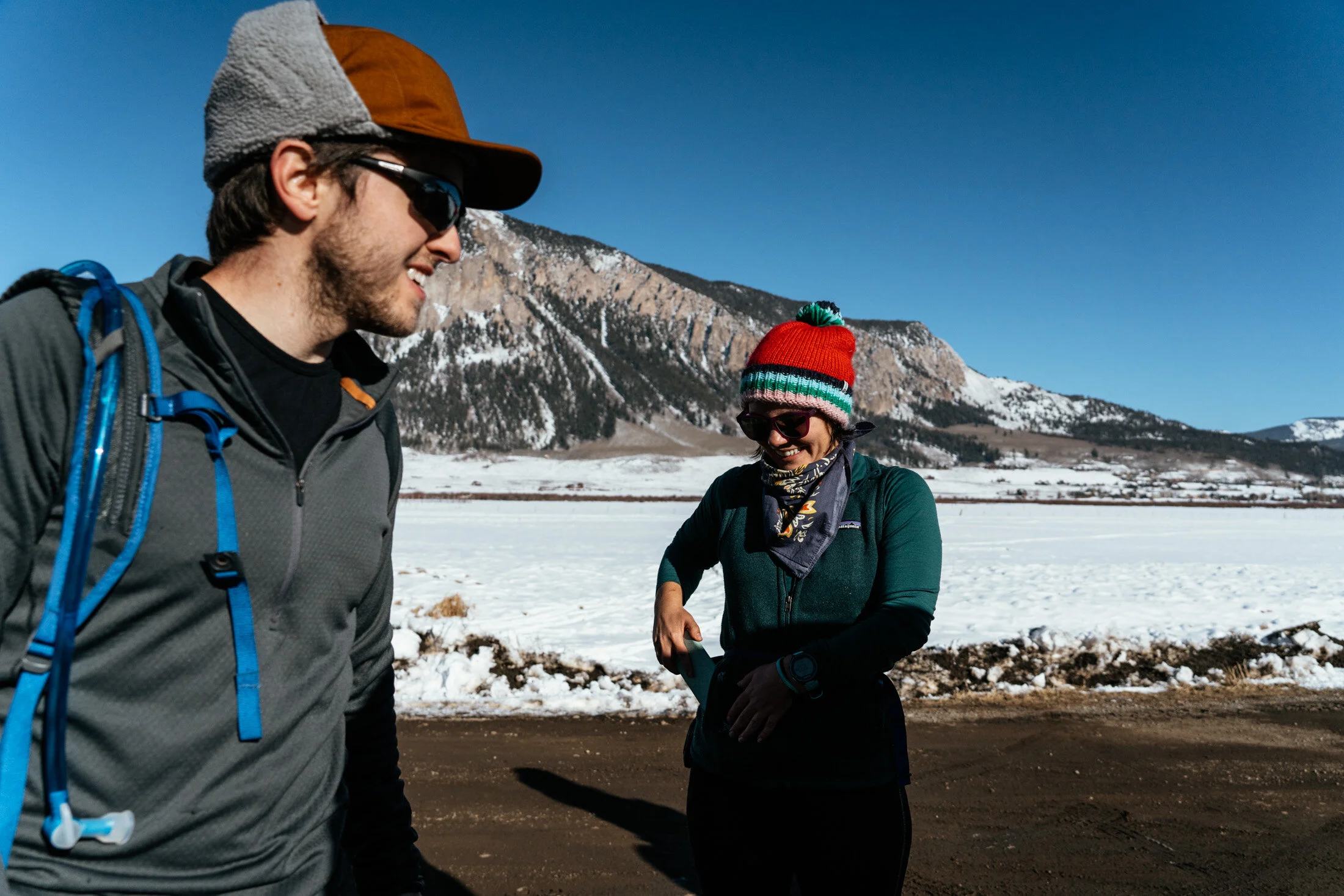 Two people in winter hiking gear smiling and talking outdoors with snow-covered mountains and blue sky in the background.