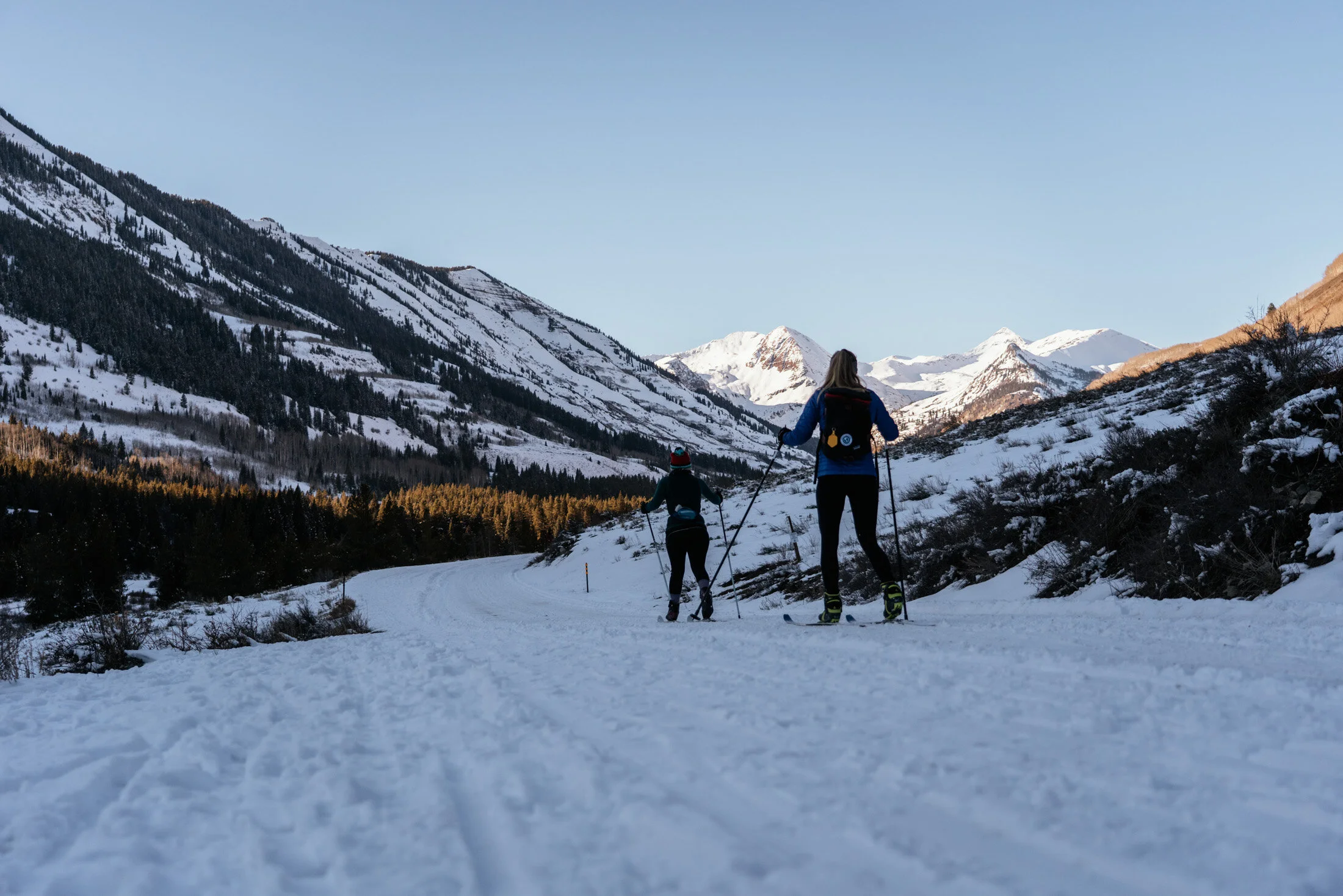 Two people cross-country skiing on a snow-covered trail in a mountainous area with snow-capped peaks and forested slopes.