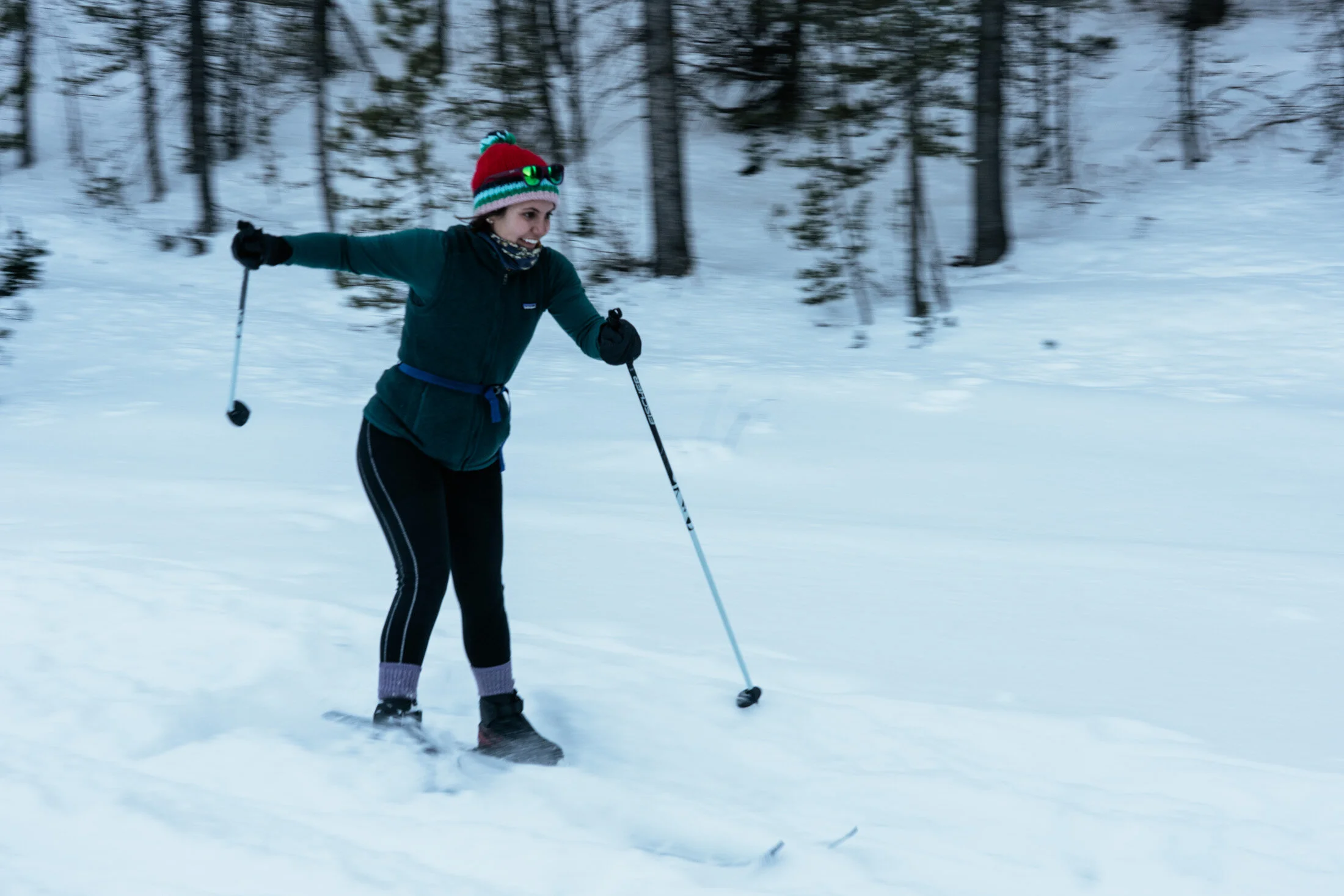 A woman cross-country skiing on snow-covered terrain with trees in the background, wearing a red hat with a pom-pom, sunglasses, and winter clothing.