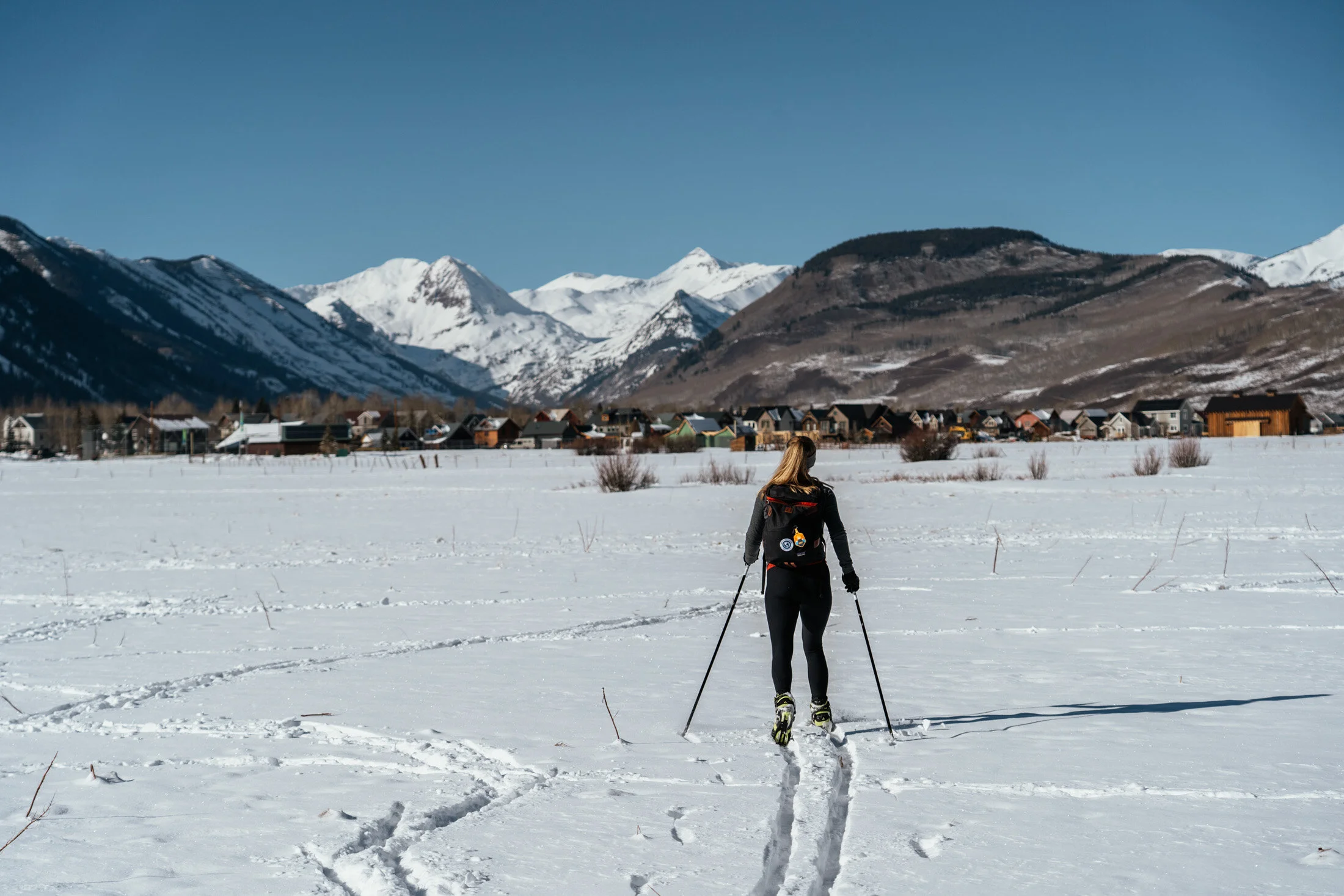A person cross-country skiing on a snow-covered field with mountains and a small town in the background.