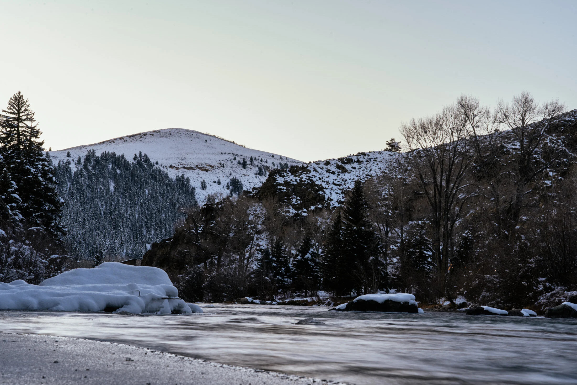 A winter landscape featuring a snowy mountain range, partially frozen river in the foreground, and snow-covered trees on the hillsides.