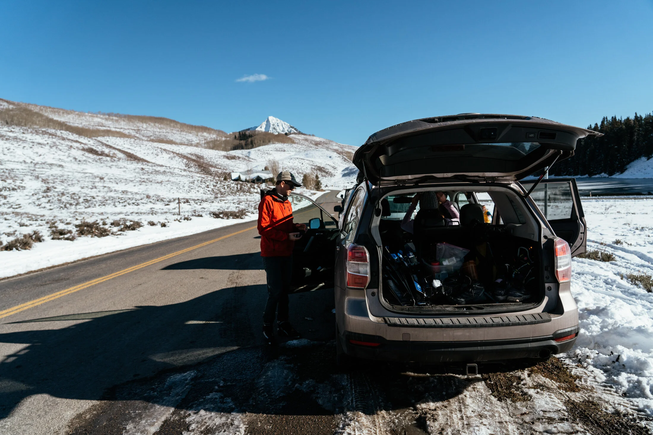 People preparing gear from an open car trunk on a snowy mountain road with snow-covered hills and a mountain peak in the background.