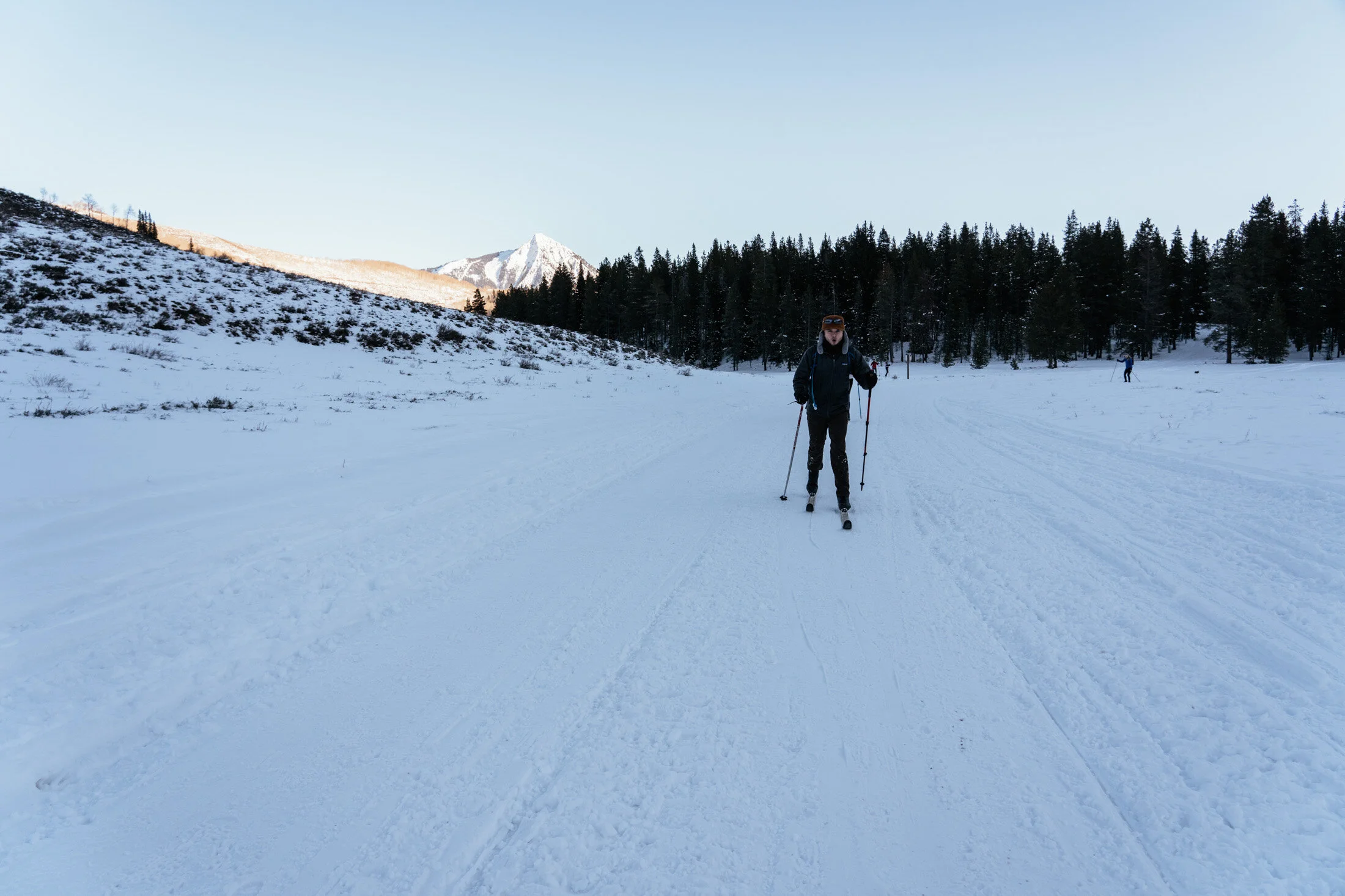 Person skiing on snow-covered trail with pine trees and mountains in the background.