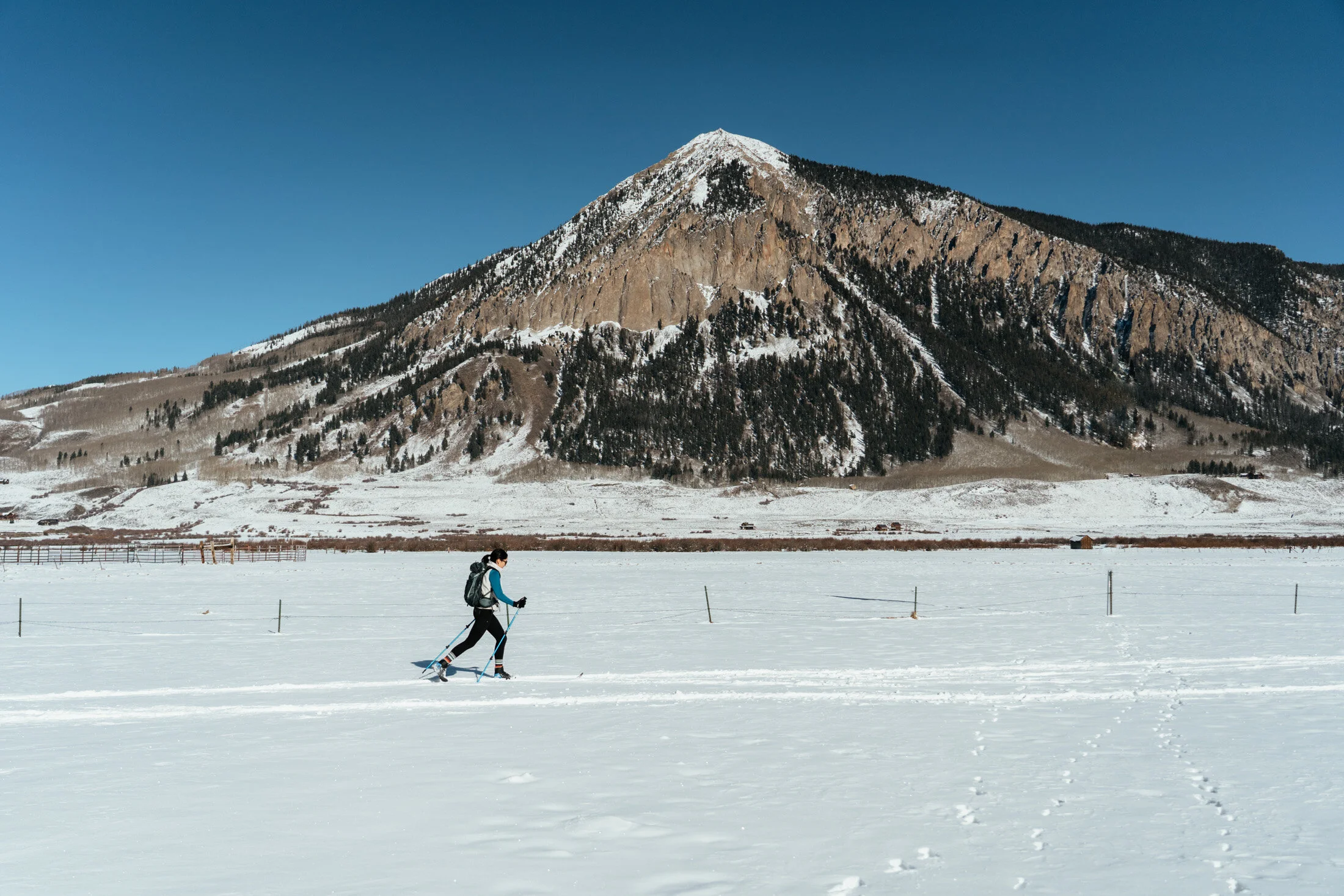 A person cross-country skiing in a snowy landscape with a mountain in the background under a clear blue sky.