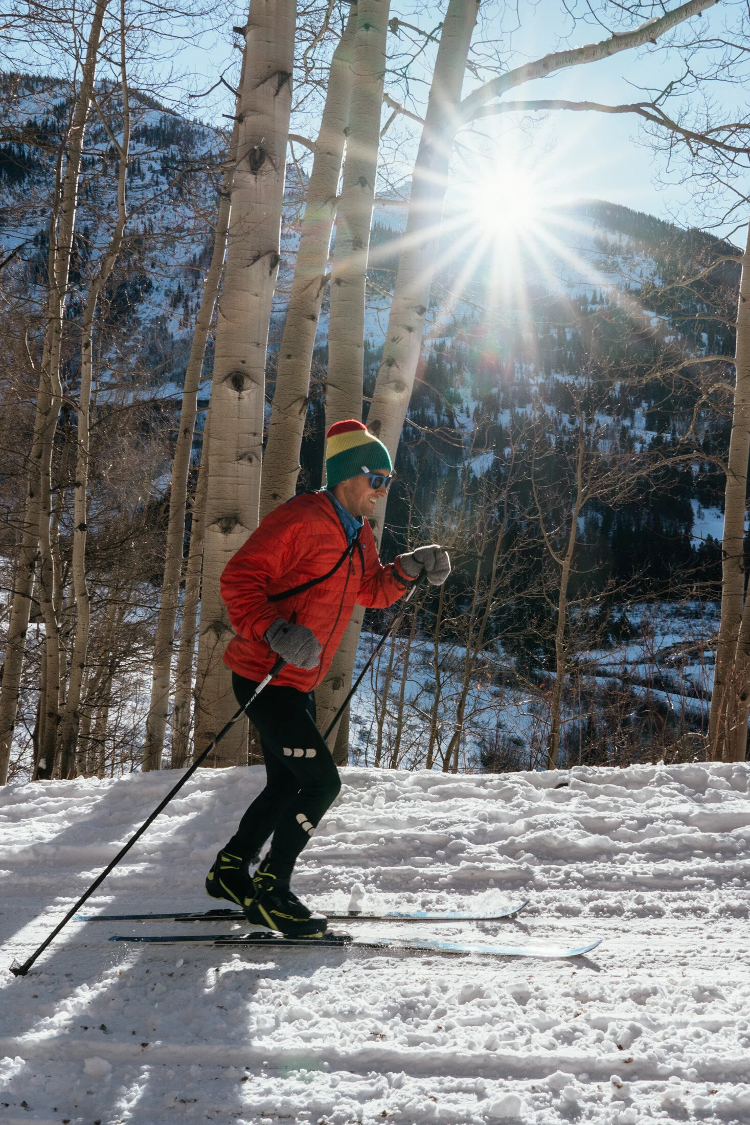 A man wearing a red jacket, gray gloves, sunglasses, and a multicolored beanie is skiing on snow for exercise during daytime with clear blue skies, dry leafless trees, and mountain scenery in the background. The sun shines brightly, creating a lens f