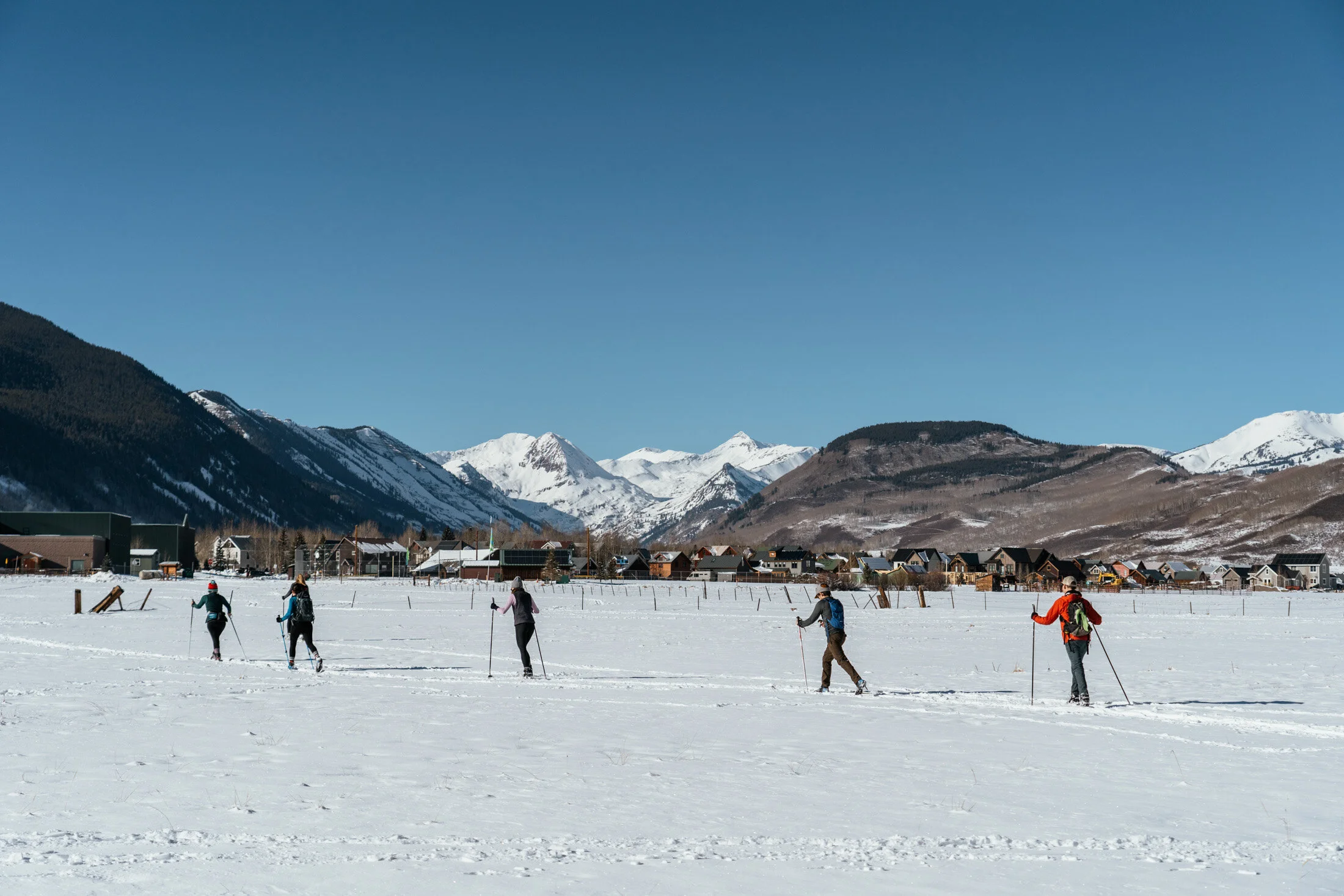People cross-country skiing in a snow-covered field with mountains and a small town in the background.