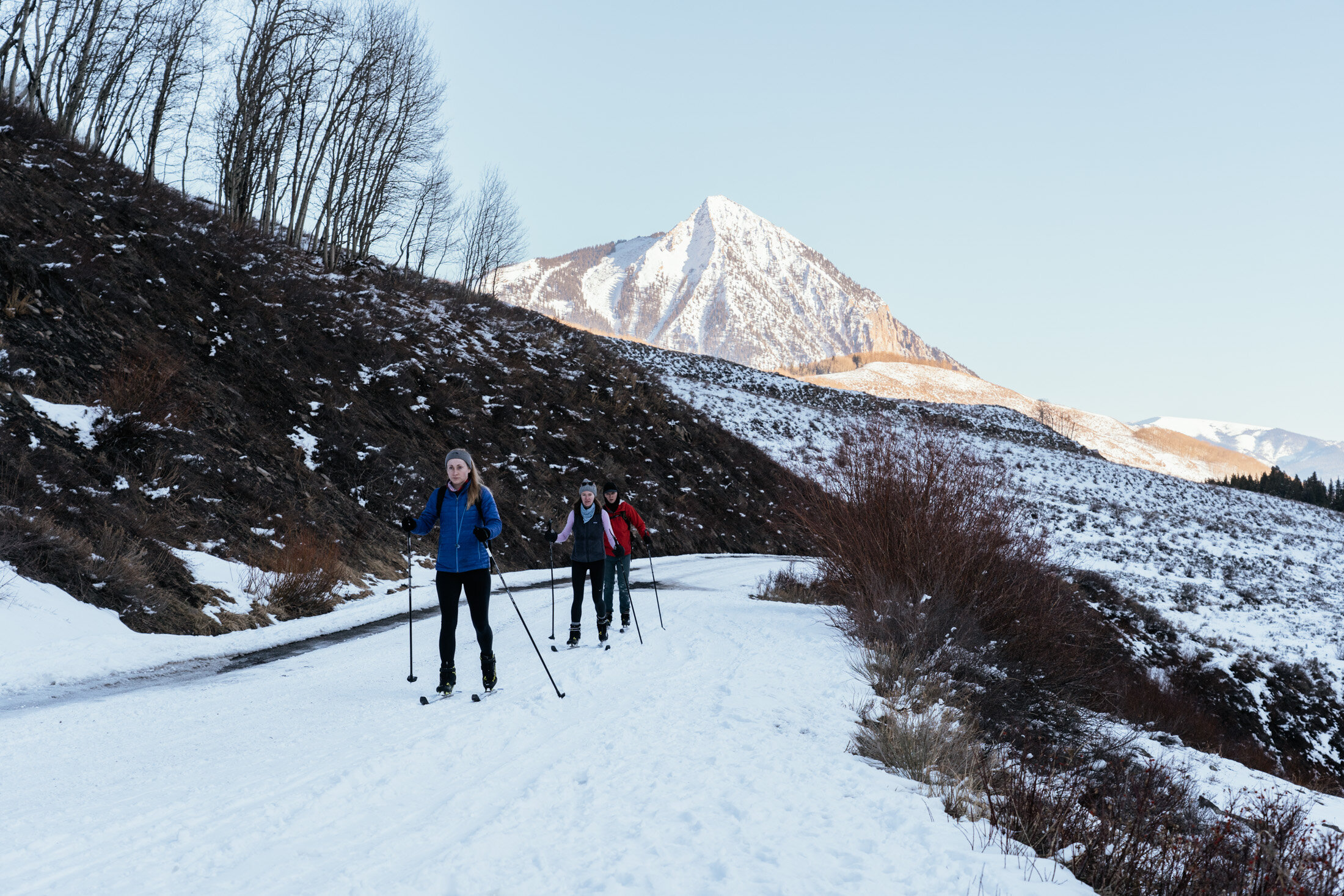 Three people cross-country skiing on a snowy trail in a mountain landscape with snow-capped peaks and leafless trees.