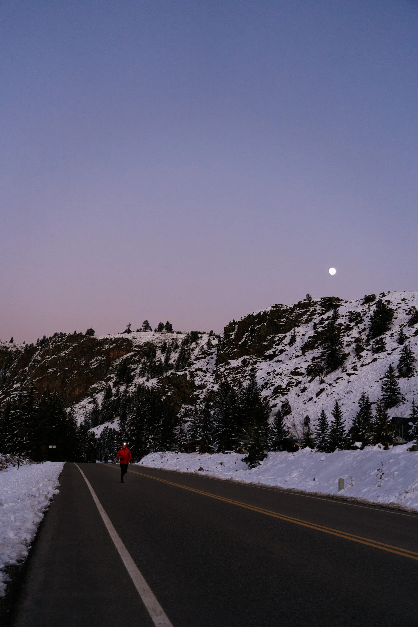 A person running on a snowy mountain road during dusk, with a mountainous landscape and trees, and the moon visible in the sky.