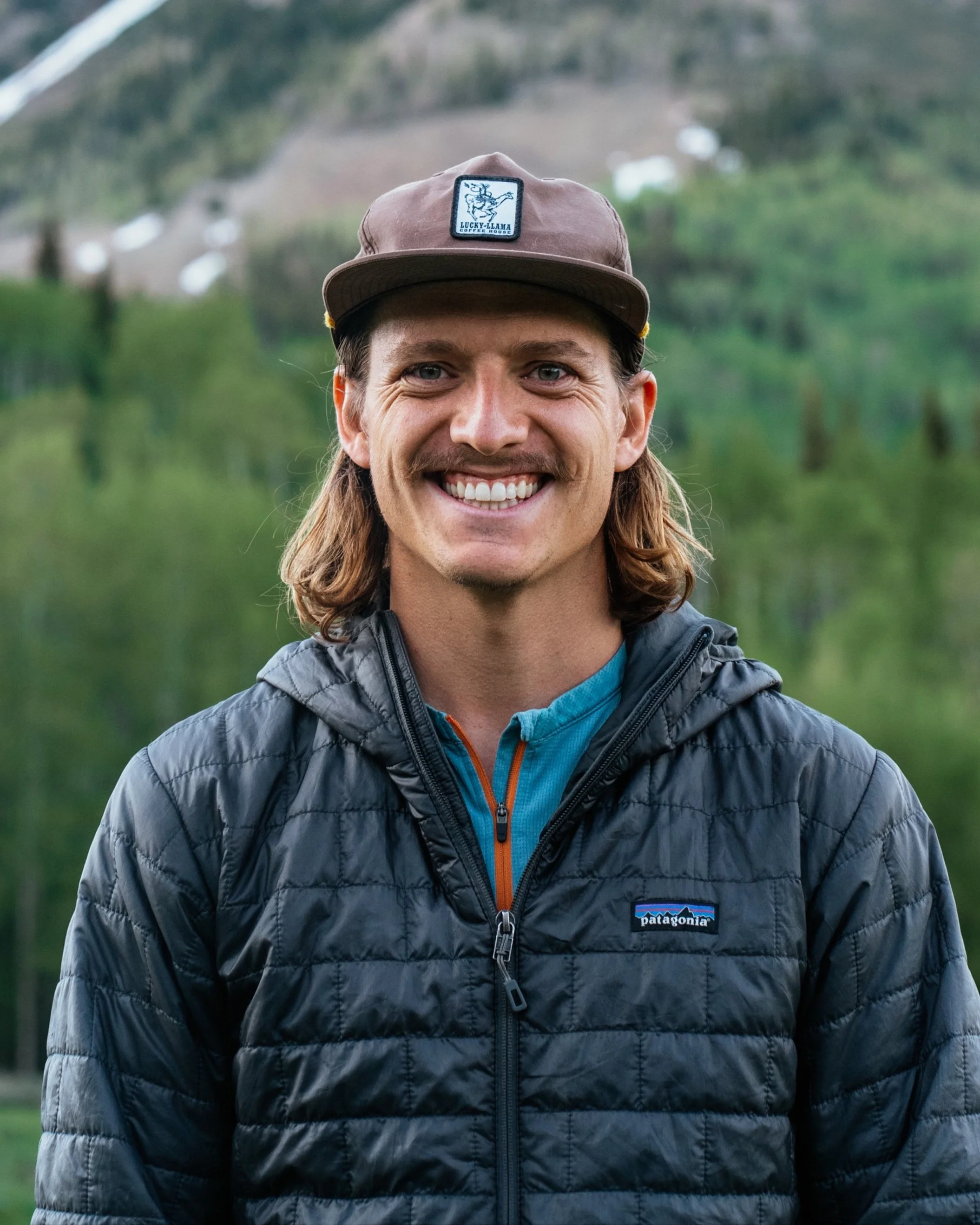 A smiling man wearing a brown Patagonia quilted jacket and a brown cap outdoors with a green mountainous landscape in the background.
