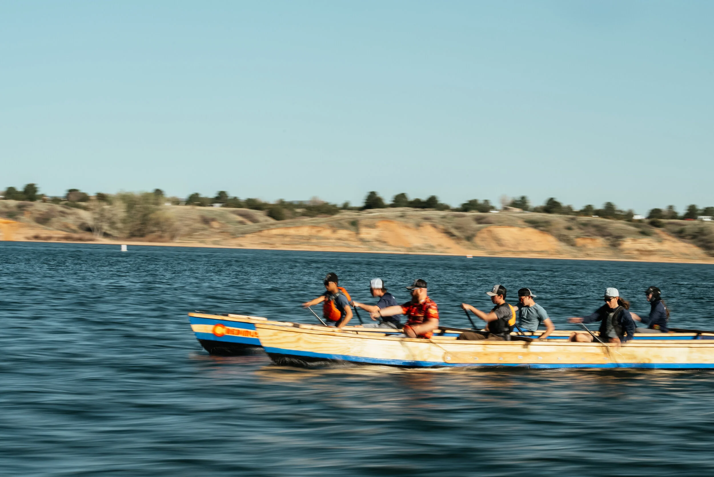 A group of eight people in a long wooden boat on a lake, with rocky hills and sparse vegetation in the background, under a clear blue sky.