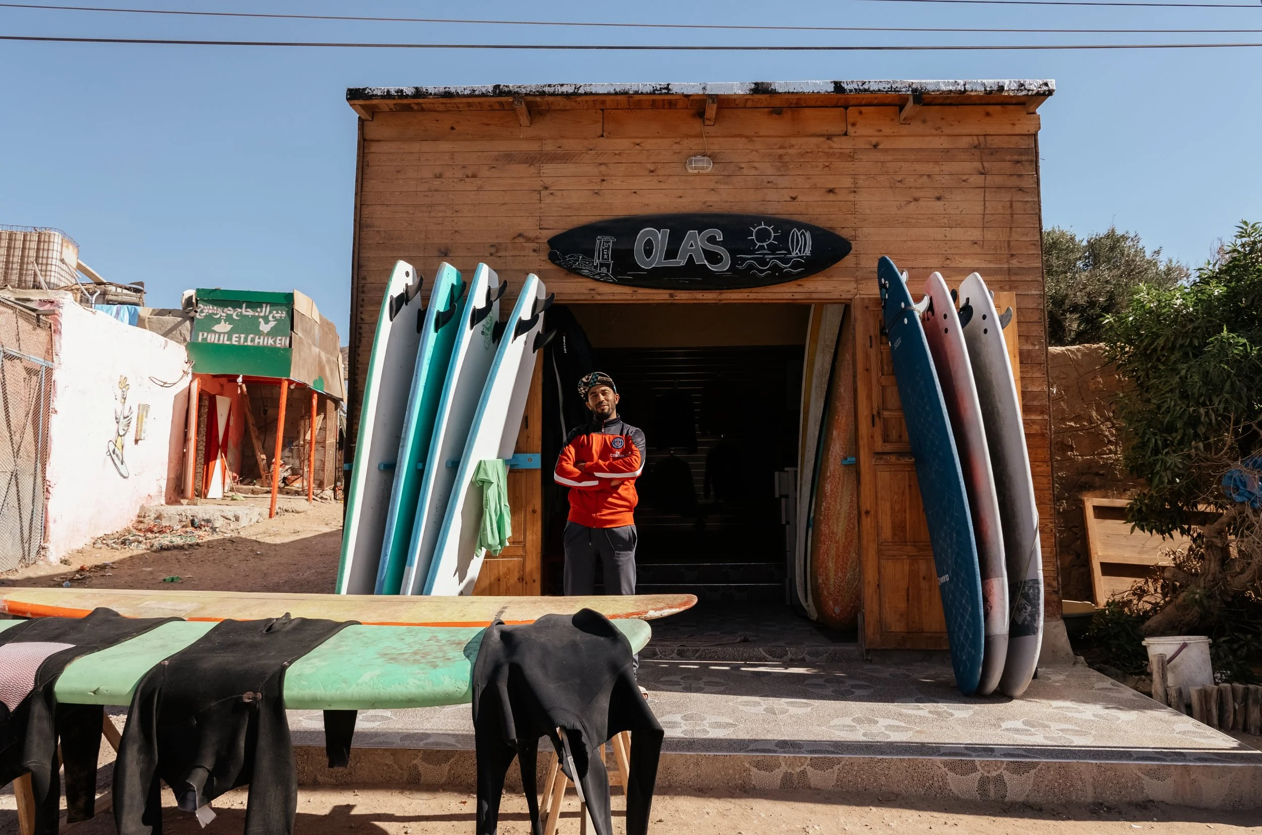 A surf shop with surfboards of various sizes and colors displayed outside. A man stands in front of the shop wearing a red jacket. The shop has a wooden exterior with a black sign reading 'OLAS' above the entrance. In the foreground, a surfboard with