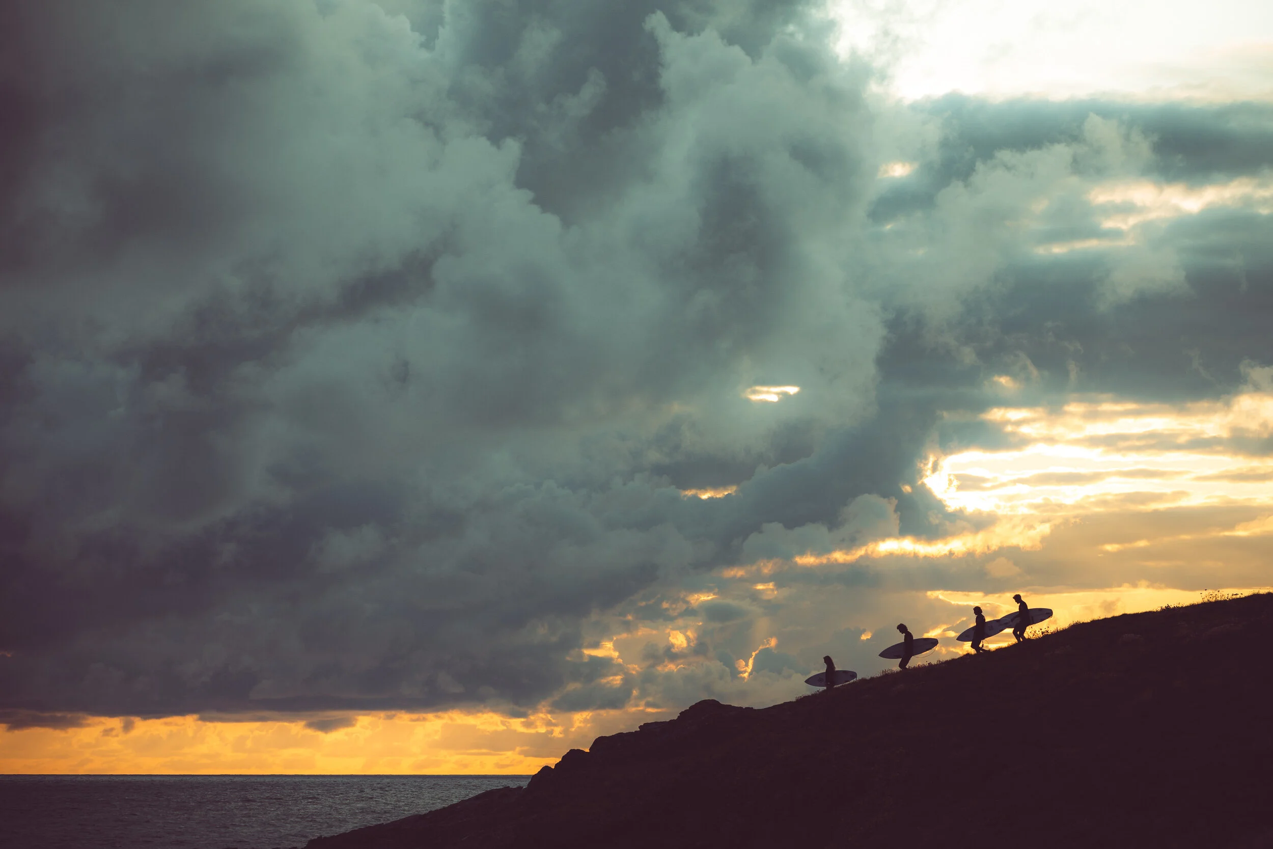Four surfers walking along a rocky shoreline with surfboards, silhouetted against a cloudy sunset sky over the ocean.