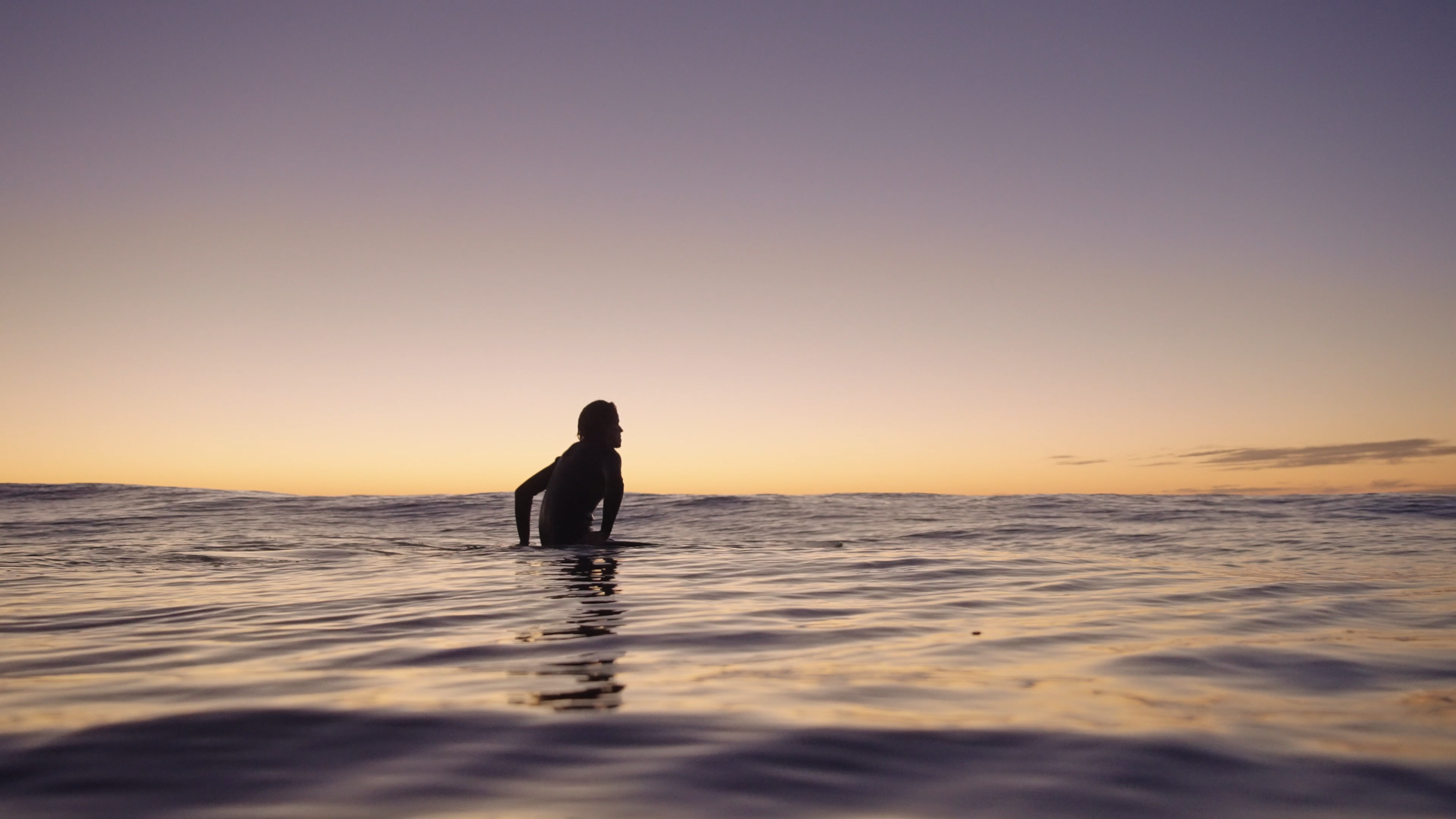 Person sitting in the shallow ocean water during sunset or sunrise with clear sky and horizon.