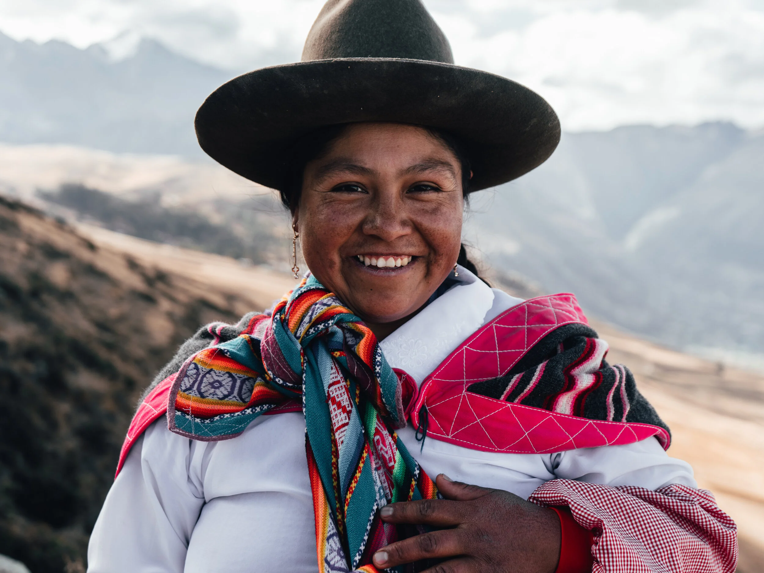A woman from the Andean region wearing traditional colorful clothing, including a wide-brimmed hat and a scarf, standing outdoors with mountains in the background, smiling at the camera.