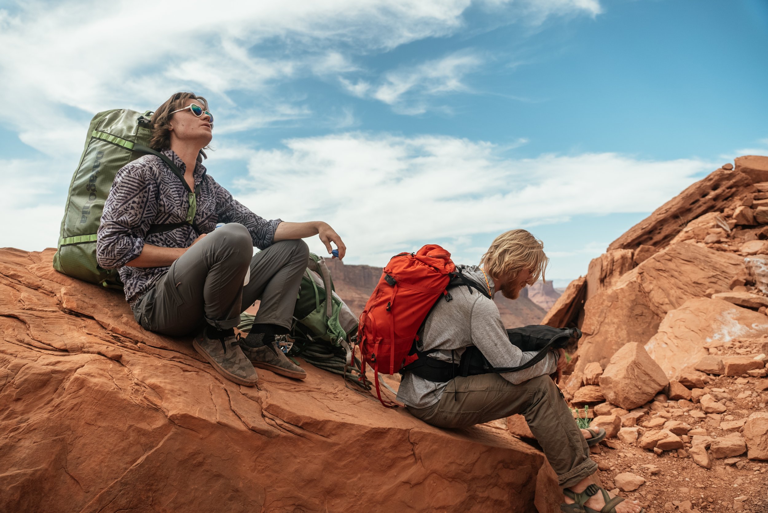 Two hikers sitting on red rocks in a desert landscape, with one reading a map and the other looking into the distance, under a blue sky with clouds.