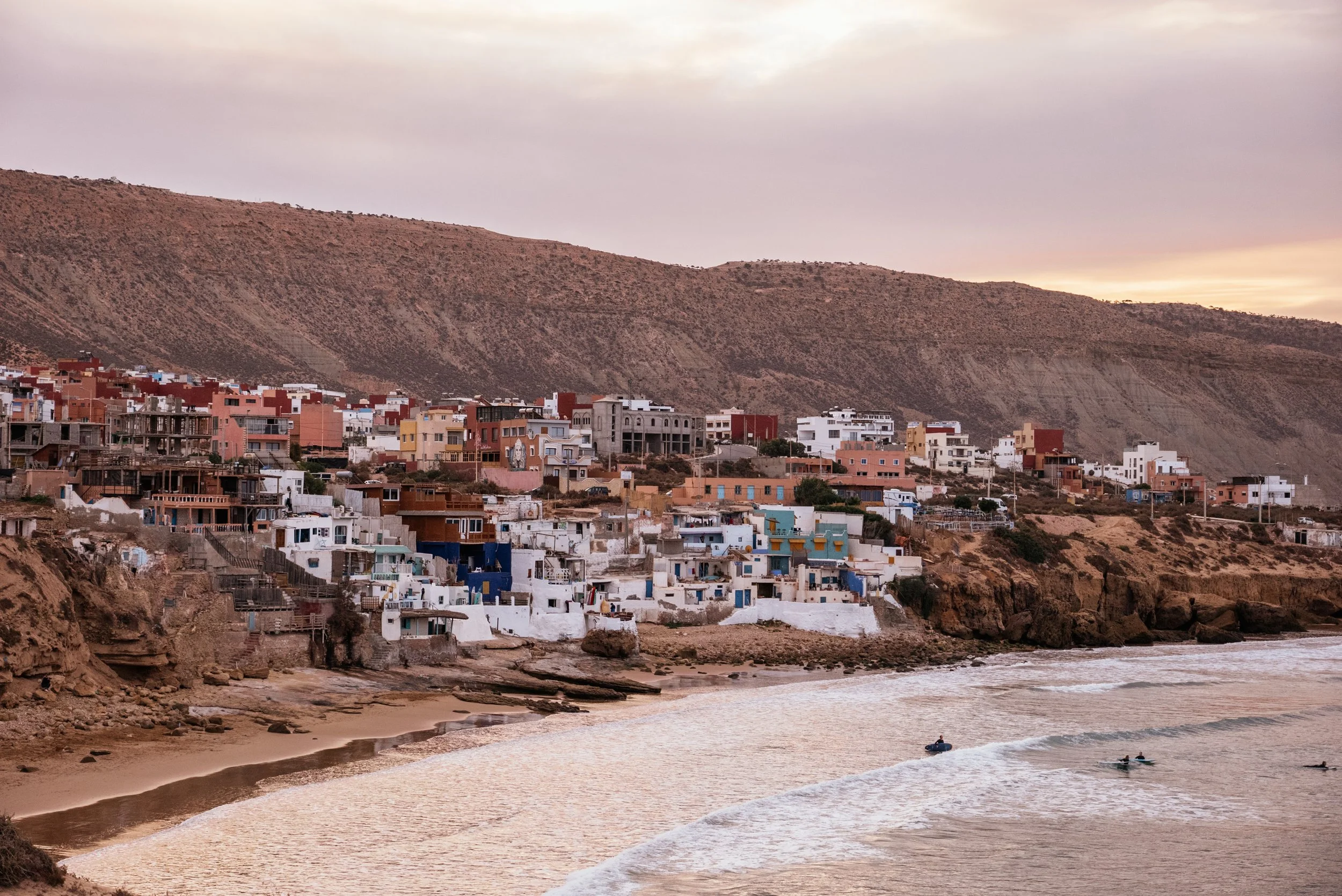 Coastal hillside town with colorful houses overlooking the beach at sunset, with boats on the water.